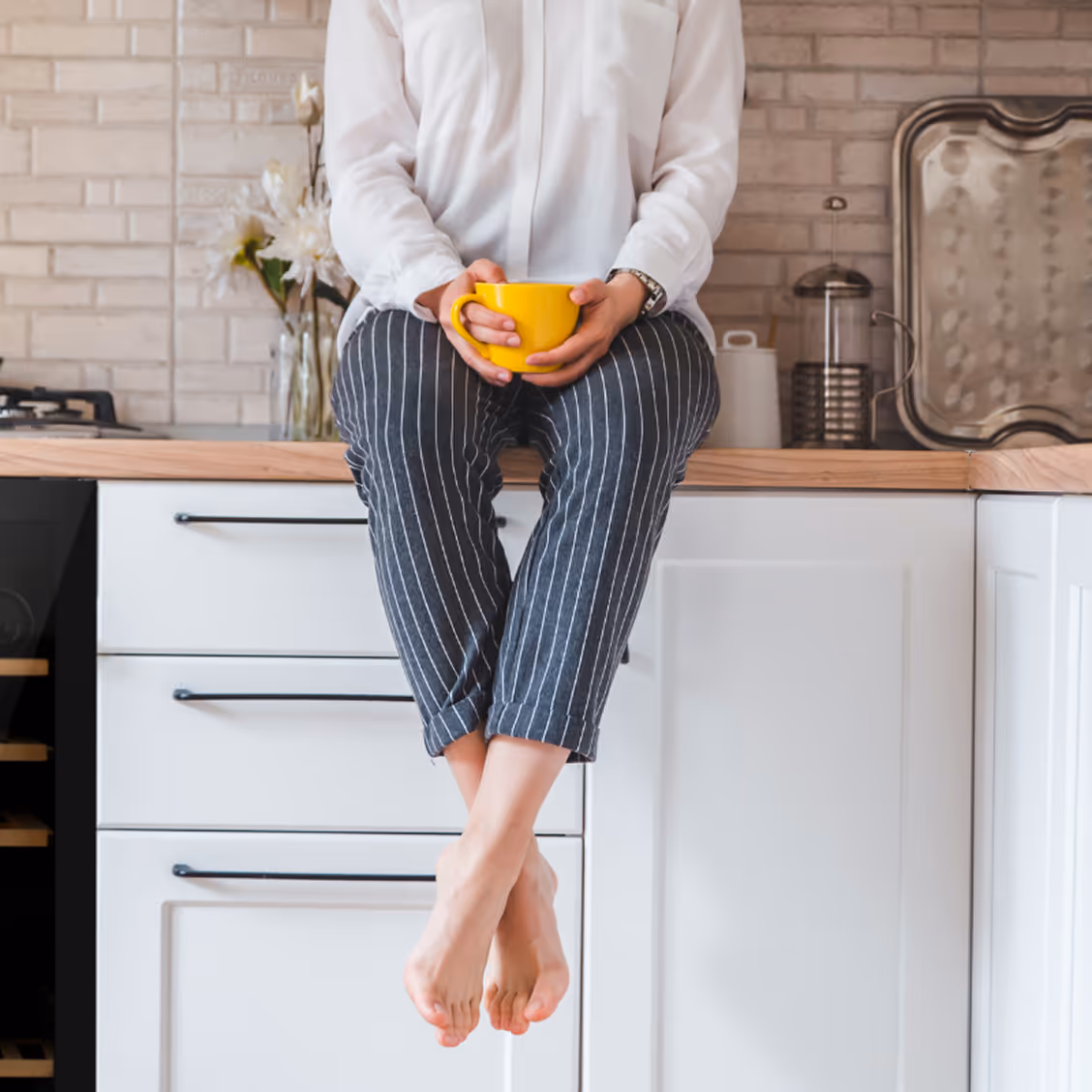 Person assise sur un plan de travail de cuisine, tenant une tasse jaune, pieds nus et pantalon rayé.