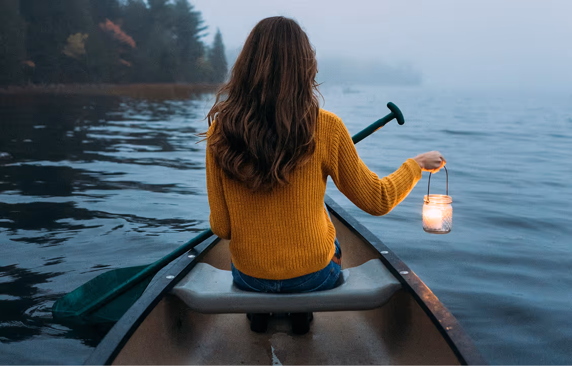 Femme dans un pull orange pagayant en canoë sur un lac calme avec une lanterne dans une main, vue de dos.