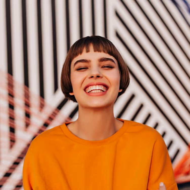 Jeune femme souriante avec une coupe au carré et un pull orange devant un mur à motifs géométriques noirs et blancs.