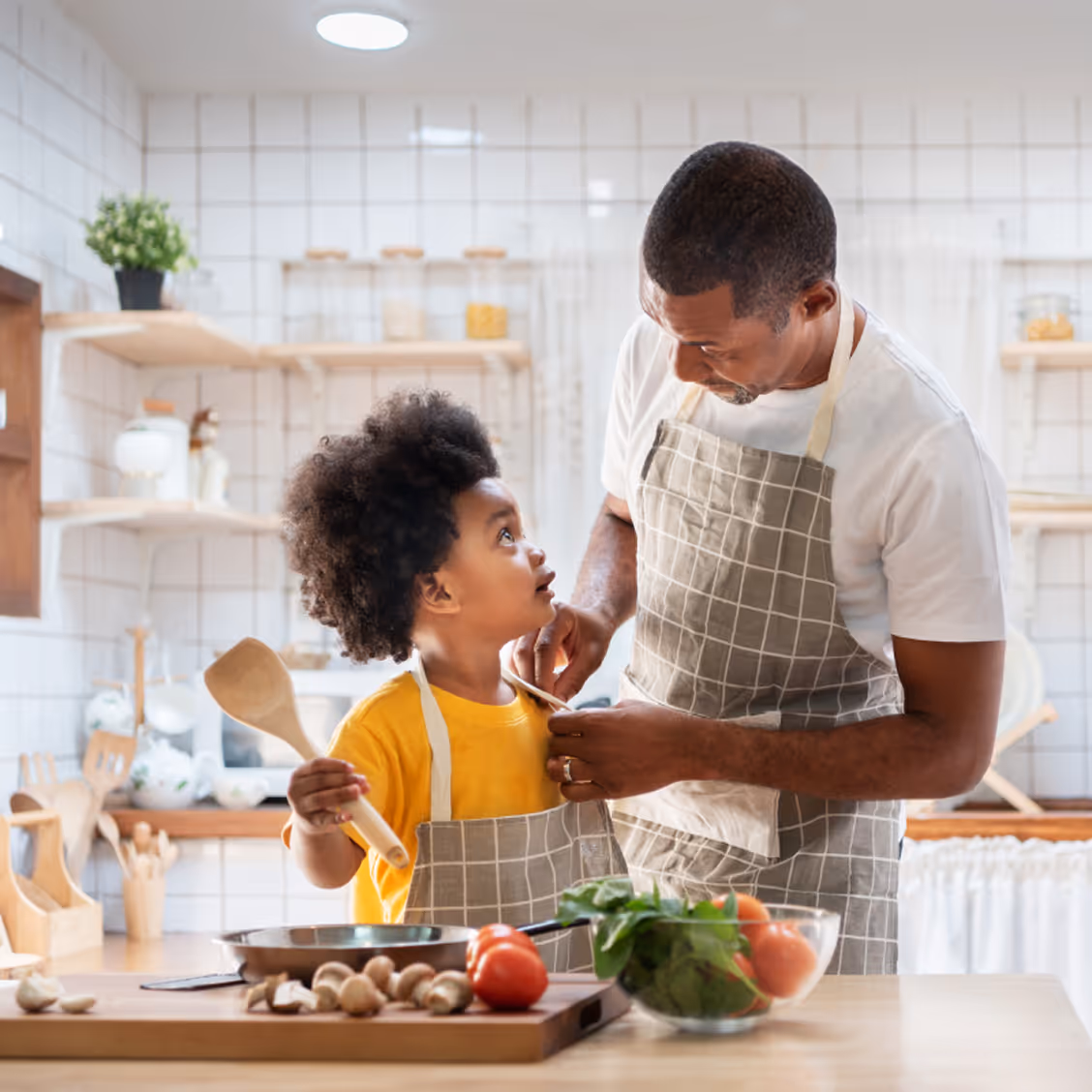 Un père attache le tablier de son enfant qui tient une cuillère en bois dans une cuisine avec des légumes frais sur le comptoir.