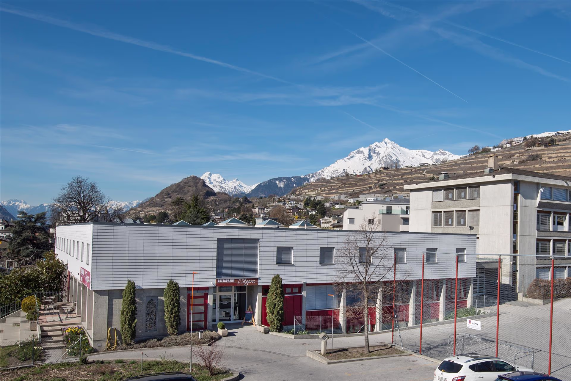 Bâtiment moderne avec entrée de restaurant, parkings, arbres, collines en terrasses et montagnes enneigées sous un ciel bleu.