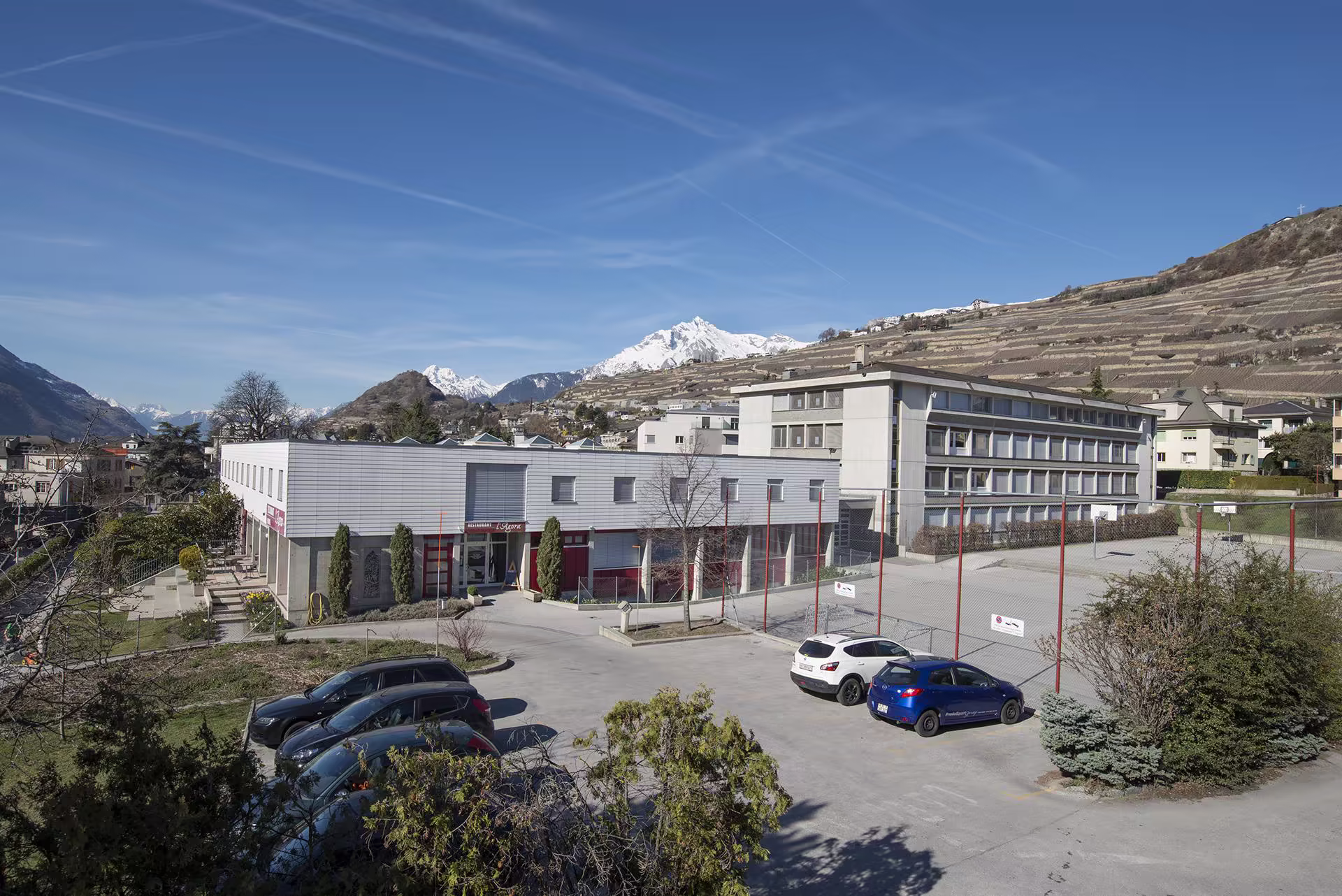 Bâtiment contemporain avec parking et voitures, terrain de sport grillagé, vignobles en terrasses et montagnes enneigées en arrière-plan sous un ciel bleu.