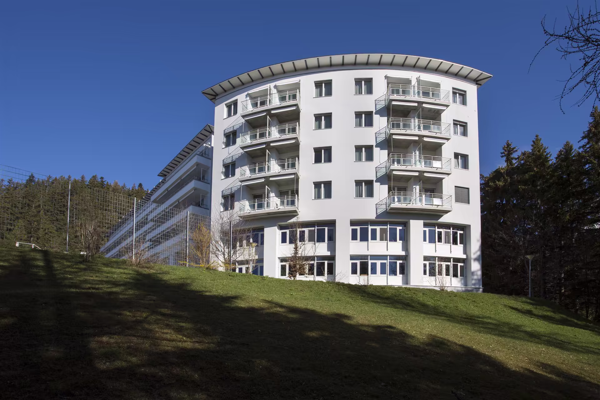 Bâtiment blanc moderne avec plusieurs balcons, situé sur une pelouse en pente, entouré d’arbres sous un ciel bleu sans nuages.
