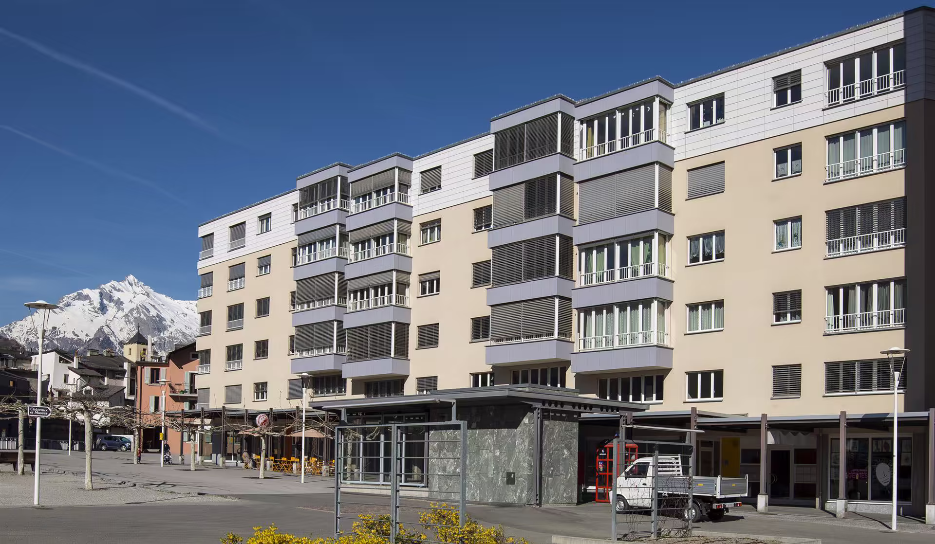 Immeuble beige et blanc avec balcons vitrés dans un quartier urbain, montagne enneigée en arrière-plan sous ciel bleu clair.