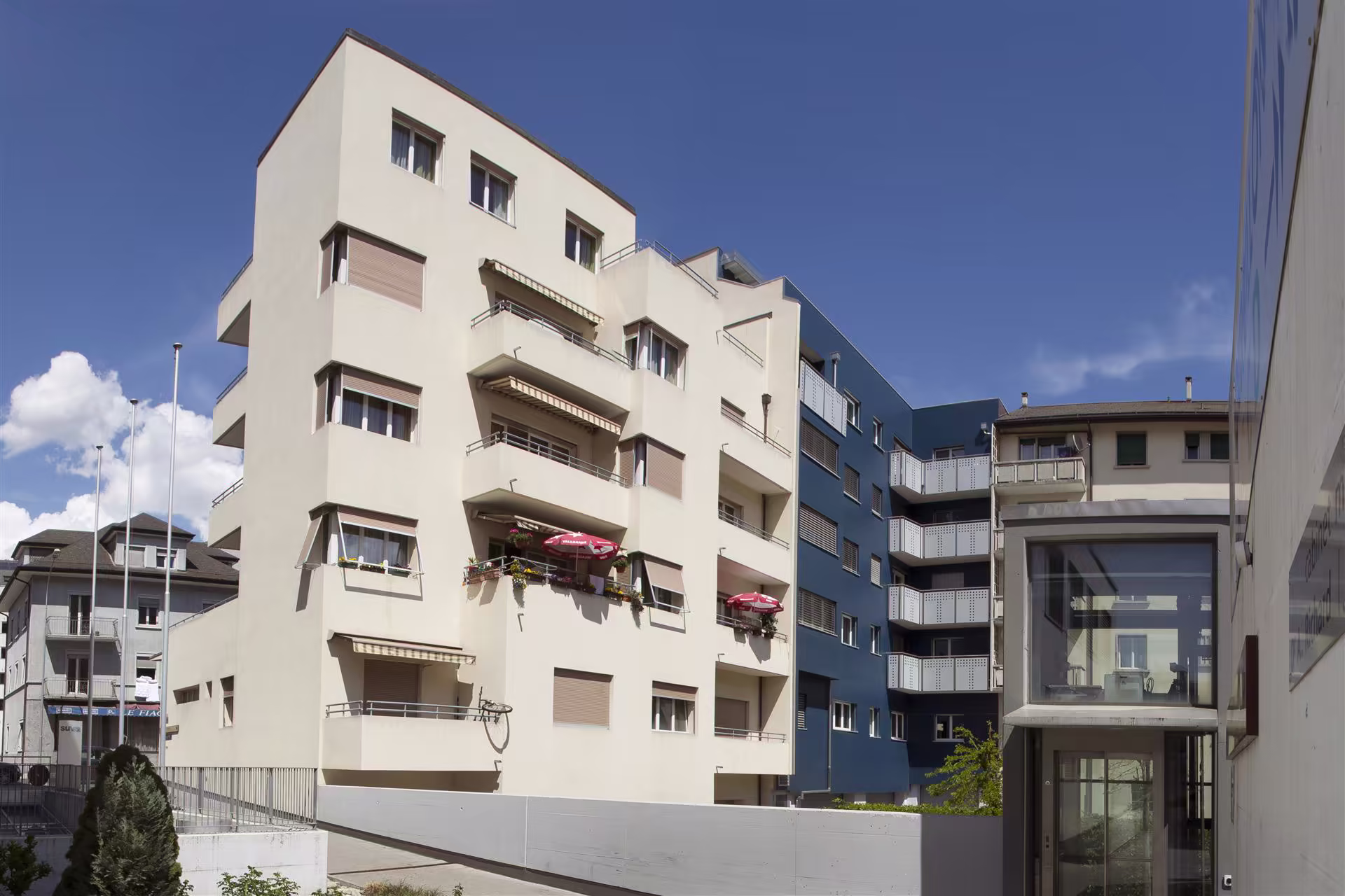 Bâtiment résidentiel moderne beige et bleu avec balcons, stores, parasols rouges, situé sous un ciel bleu clair avec quelques nuages blancs.