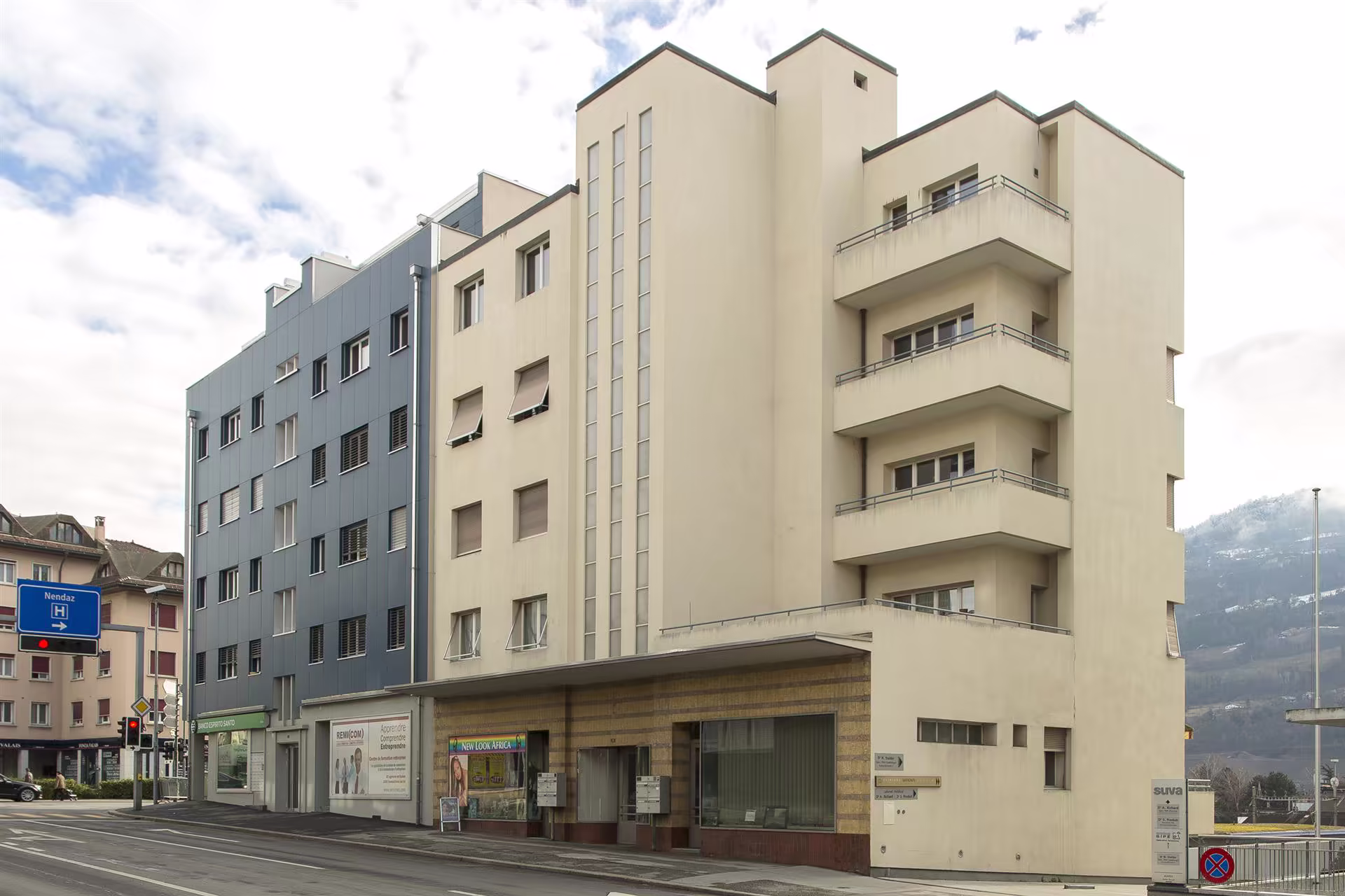 Bâtiment beige et gris de plusieurs étages avec balcons et commerces en rez-de-chaussée, situé à un coin de rue, sous ciel nuageux.