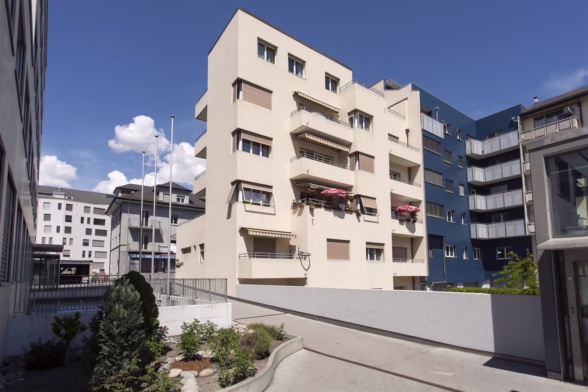 Ensemble de bâtiments modernes d'appartements lumineux avec balcons, stores, parasols rouges, et petites plantations sous un ciel bleu partiellement nuageux.