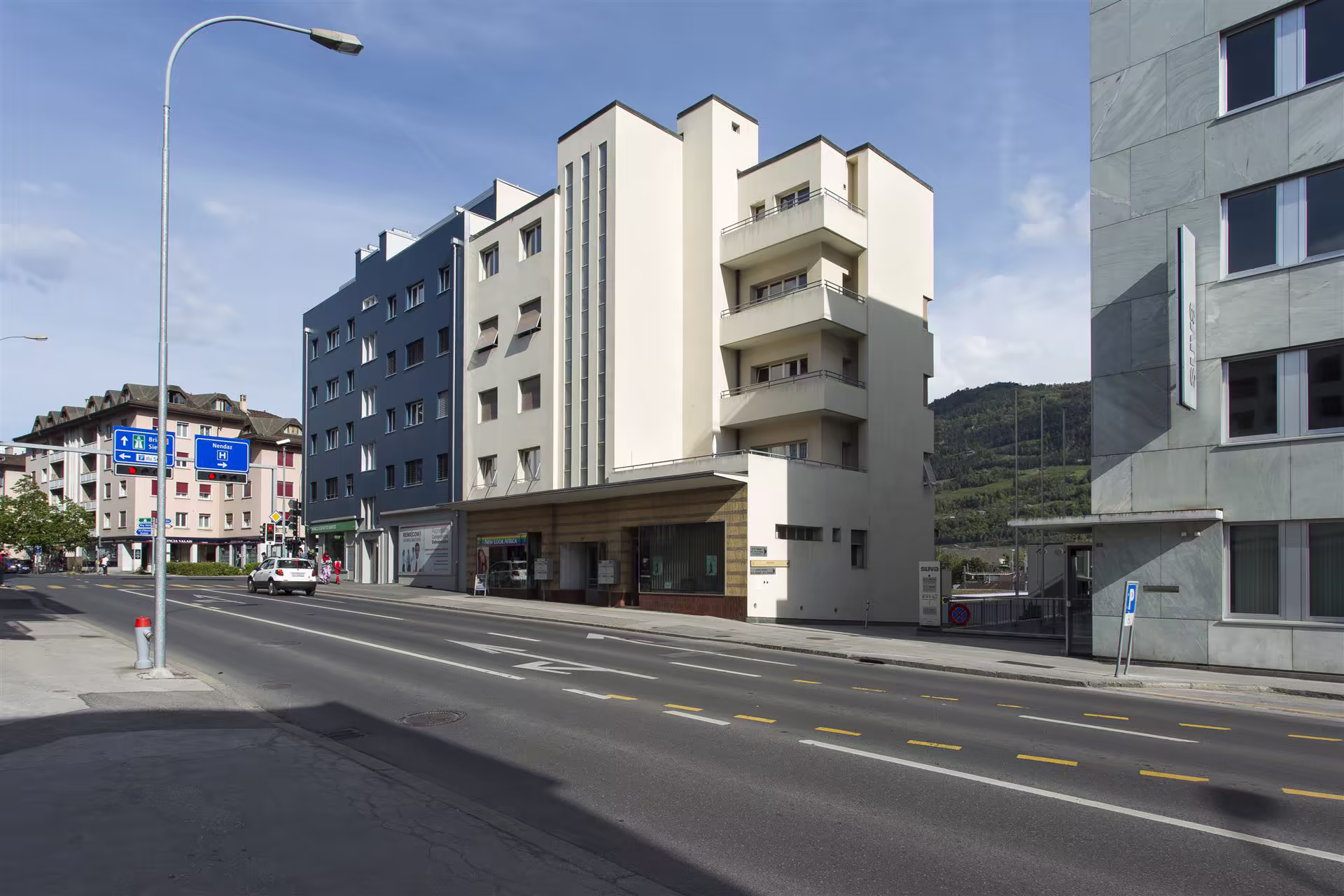 Rue urbaine avec un bâtiment blanc moderne à balcon, un immeuble gris à côté, panneaux de signalisation et voitures en circulation.