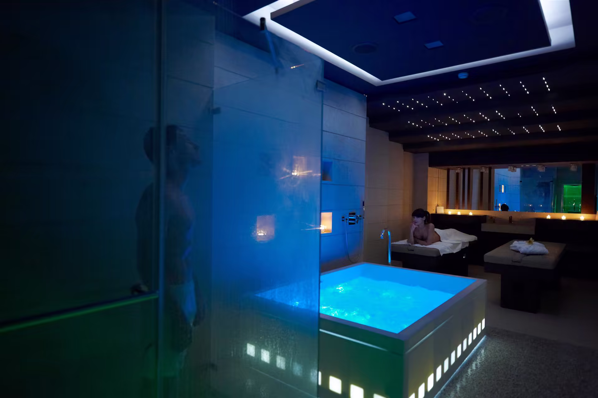 Salle de spa tamisée avec jacuzzi éclairé en bleu, homme sous une douche vitrée et femme allongée sur une table de massage, bougies allumées en arrière-plan.