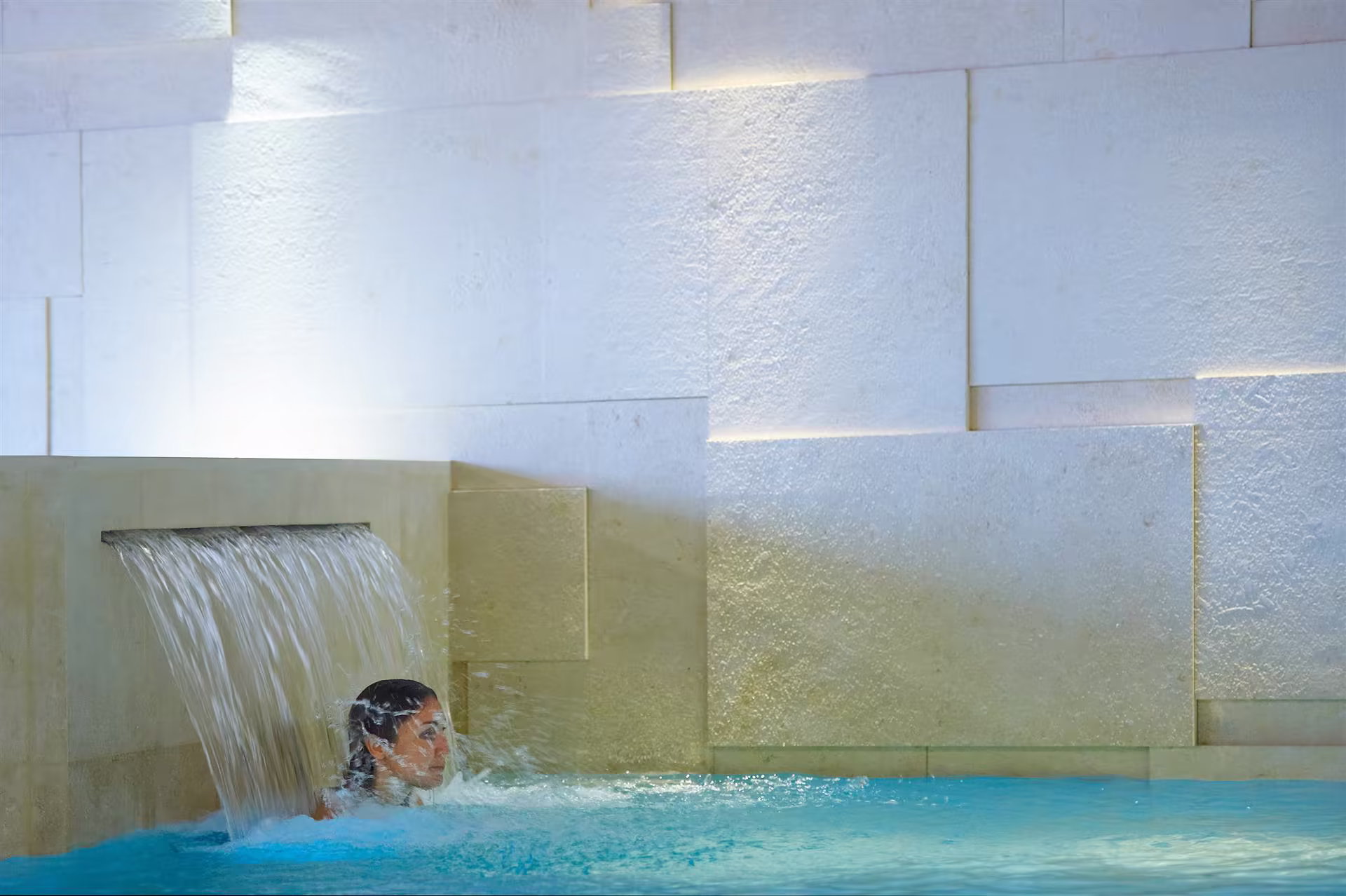 Personne sous un jet d’eau dans une piscine intérieure avec un mur de pierres blanches en arrière-plan, ambiance calme et lumineuse.