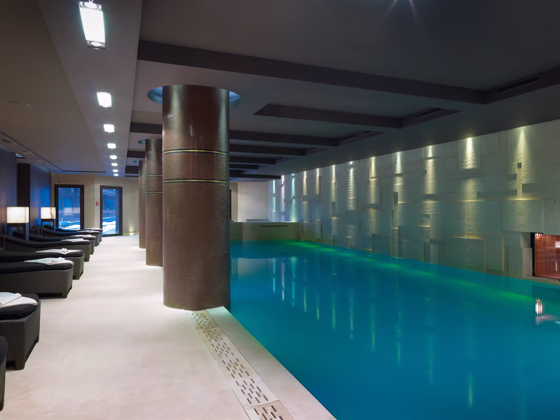 Piscine intérieure moderne avec eau bleue éclairée, colonnes recouvertes de mosaïques, mur texturé et chaises longues avec serviettes.