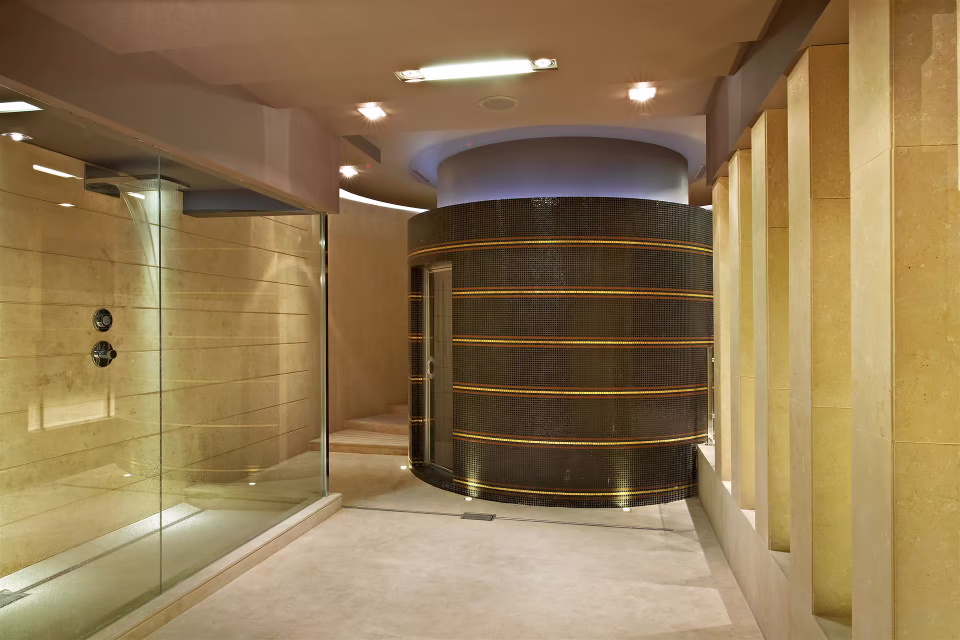 Salle de bains avec douche à l’italienne vitrée à gauche, sol beige, murs en pierre claire et paroi arrondie en mosaïque noire avec bandes lumineuses.