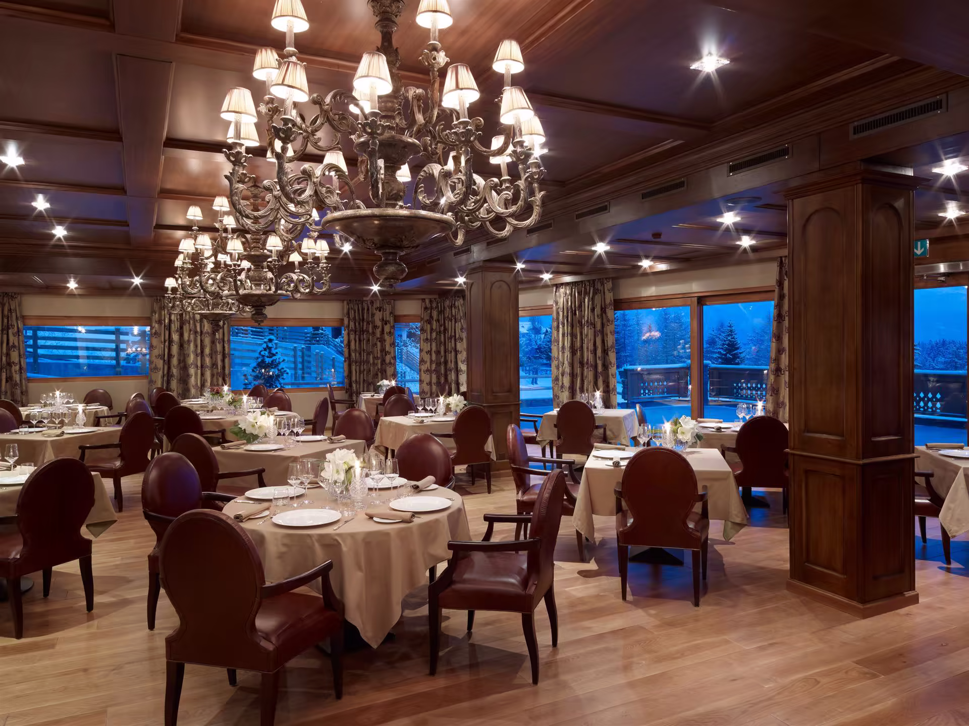 Salle de restaurant élégante avec tables nappées, chaises en cuir marron, grands lustres, murs et plafond en bois, vue sur paysage enneigé.