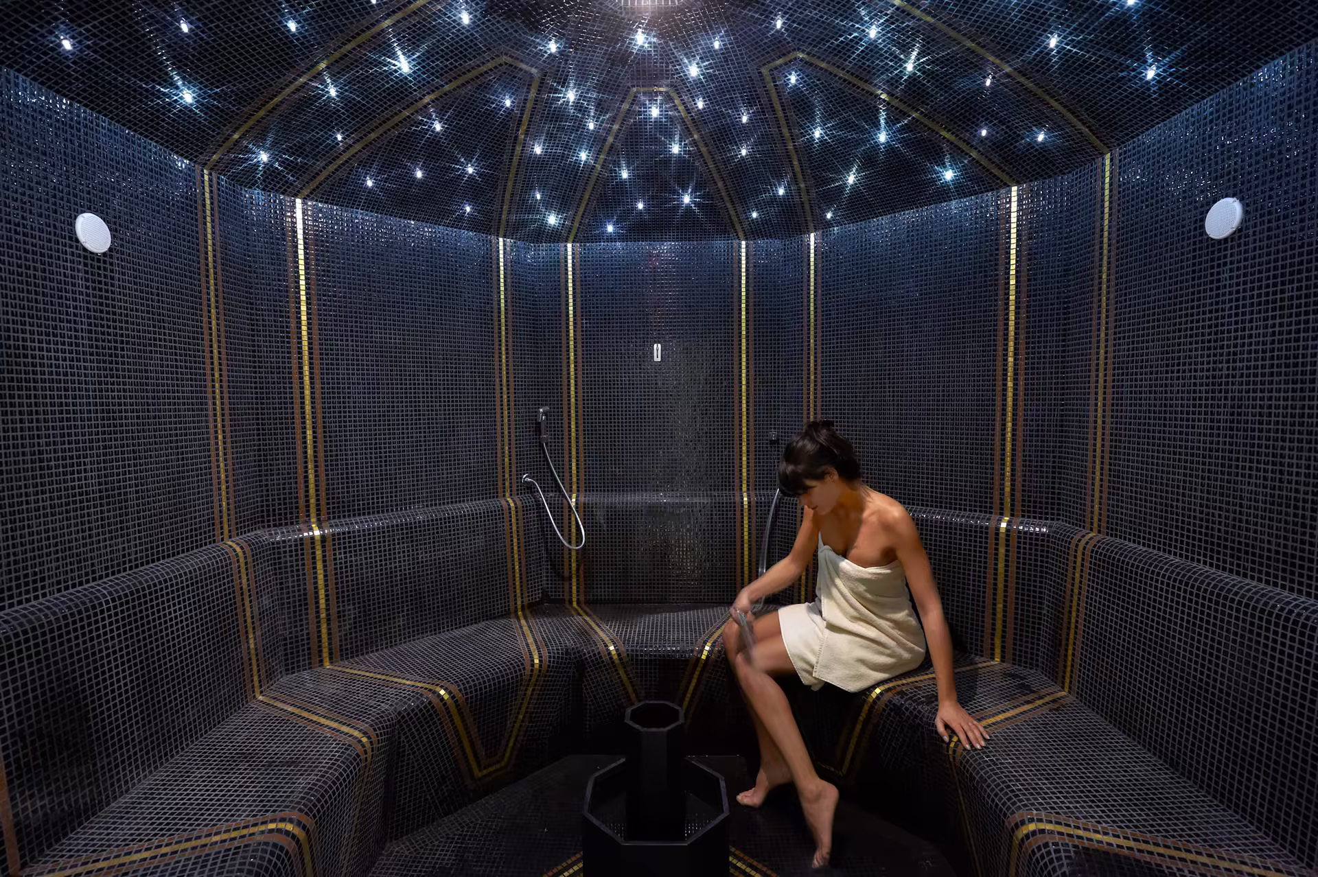 Femme en serviette blanche assise dans un hammam noir à mosaique avec plafond lumineux en étoiles, utilisant une poignée d’eau sur sa cuisse.
