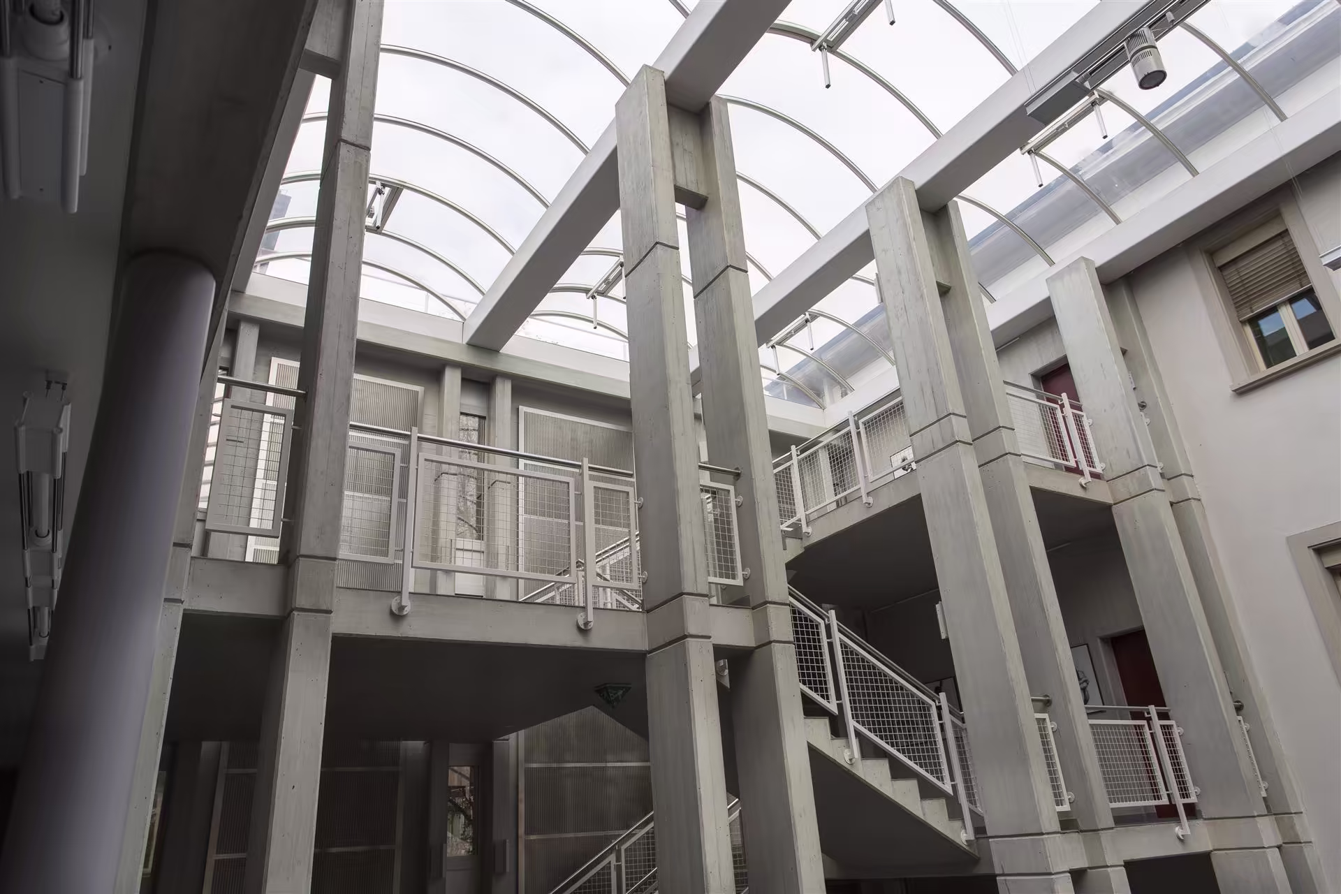 Intérieur d’un bâtiment avec grande verrière arrondie, colonnes en béton, escalier et balustrades métalliques peintes en blanc.