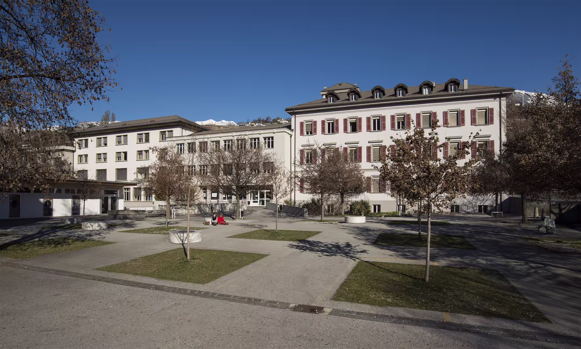 Deux personnes assises sur un banc dans une cour dégagée avec des arbres sans feuilles, entourée de deux bâtiments blancs sous un ciel bleu clair.