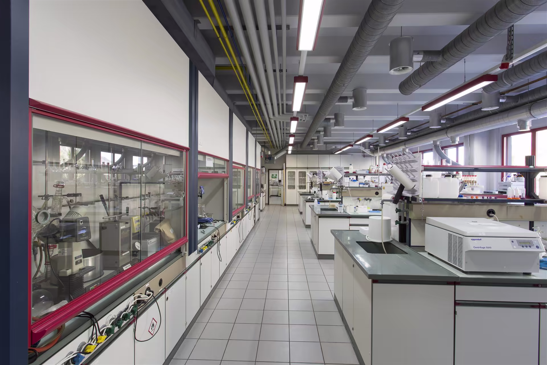 Laboratoire scientifique moderne avec paillasses, équipements de centrifugation, mobilier blanc, grandes fenêtres et conduits d'aération au plafond.