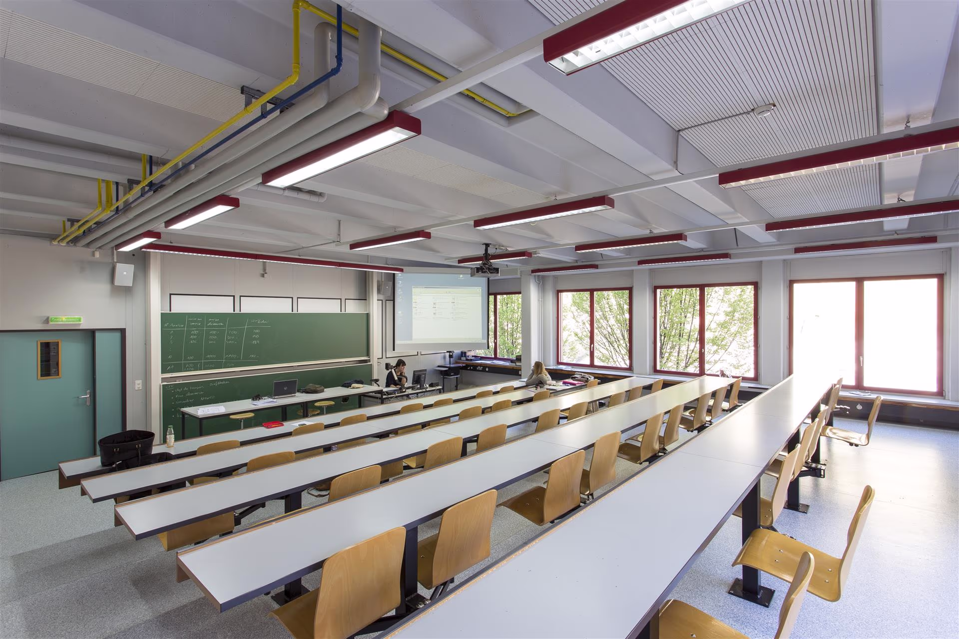 Salle de cours universitaire vide avec rangées de tables blanches et chaises en bois, tableau vert, fenêtres lumineuses et deux personnes au fond.