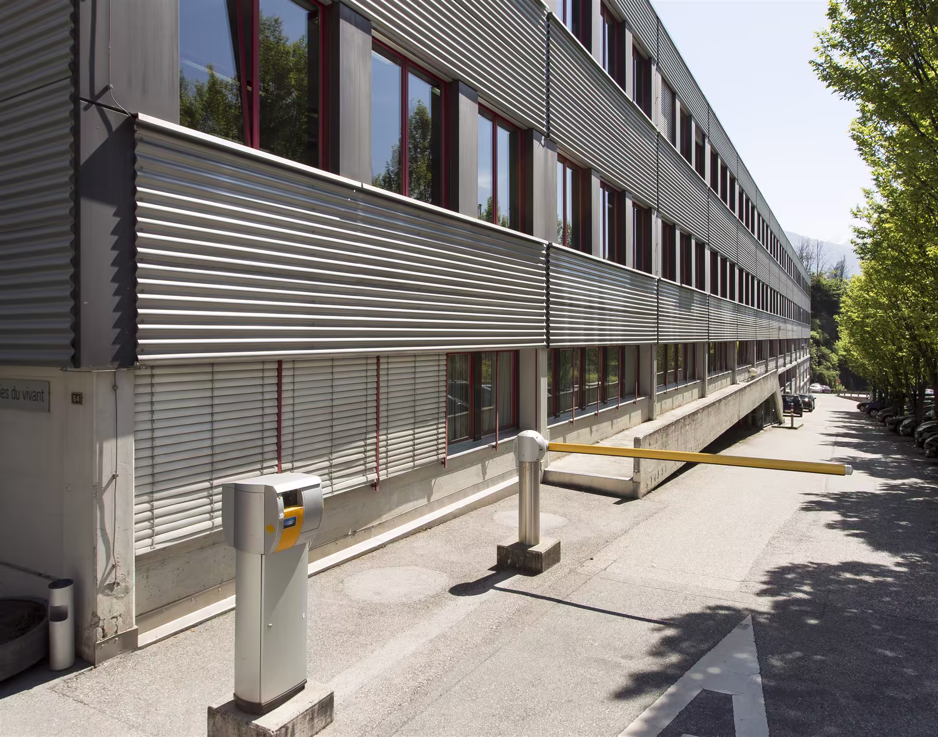 Façade moderne en métal d’un bâtiment long avec barrières automatiques jaunes et arbres alignés le long d’une route avec voitures stationnées.