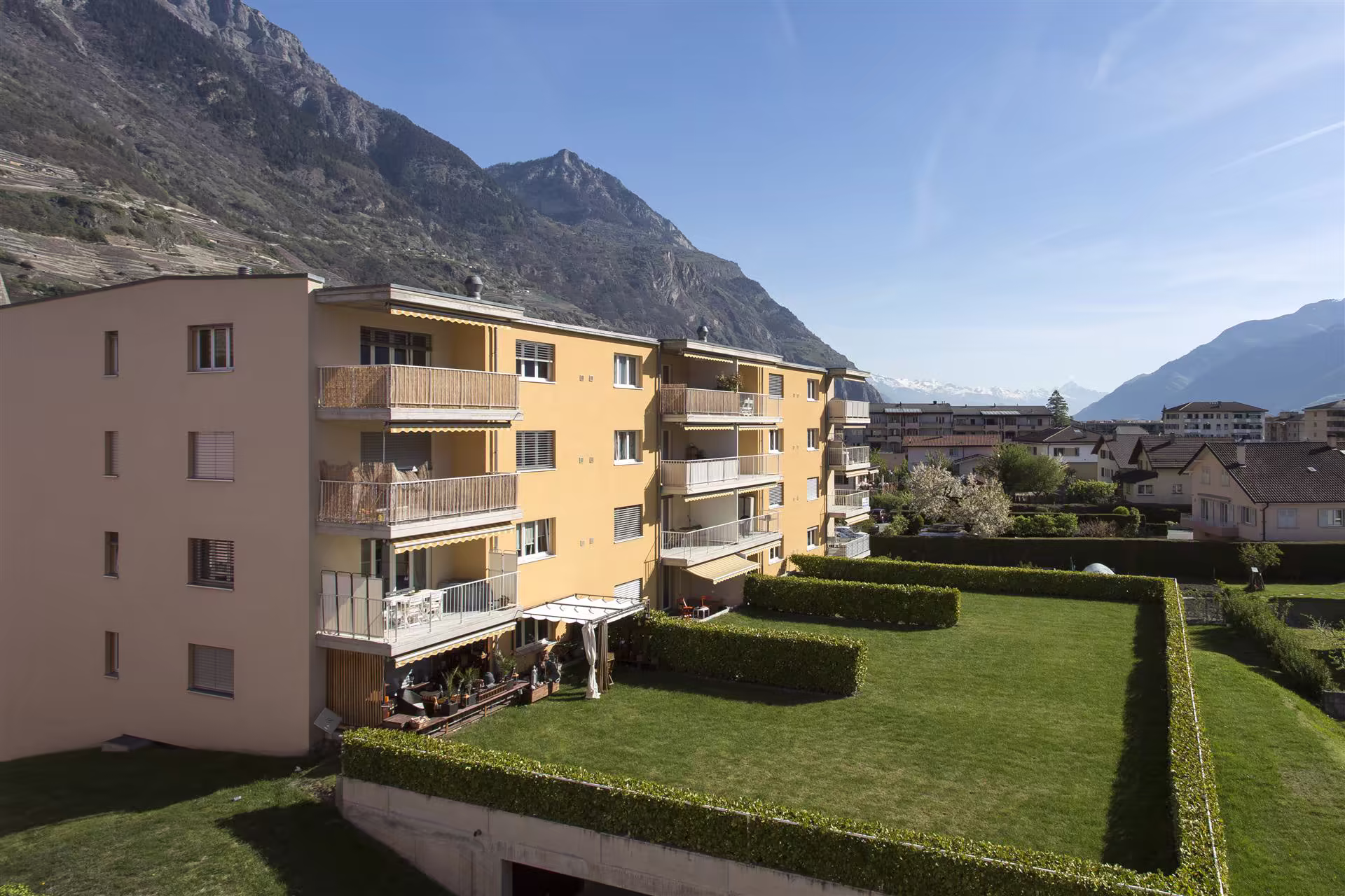 Bâtiment résidentiel jaune avec balcons, jardin bien entretenu délimité par des haies, montagnes en arrière-plan sous un ciel bleu clair.