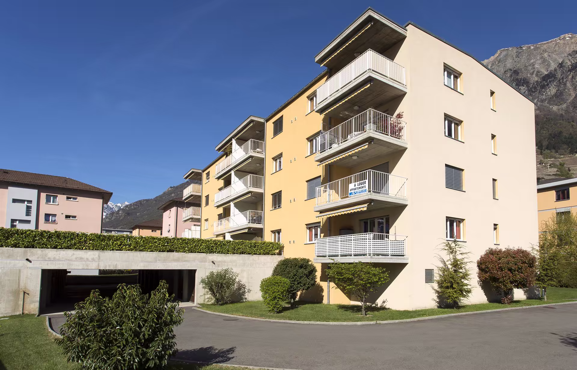 Immeuble résidentiel jaune pâle à quatre étages avec balcons, entouré d'arbustes et d'une entrée de garage, ciel bleu et montagnes en arrière-plan.