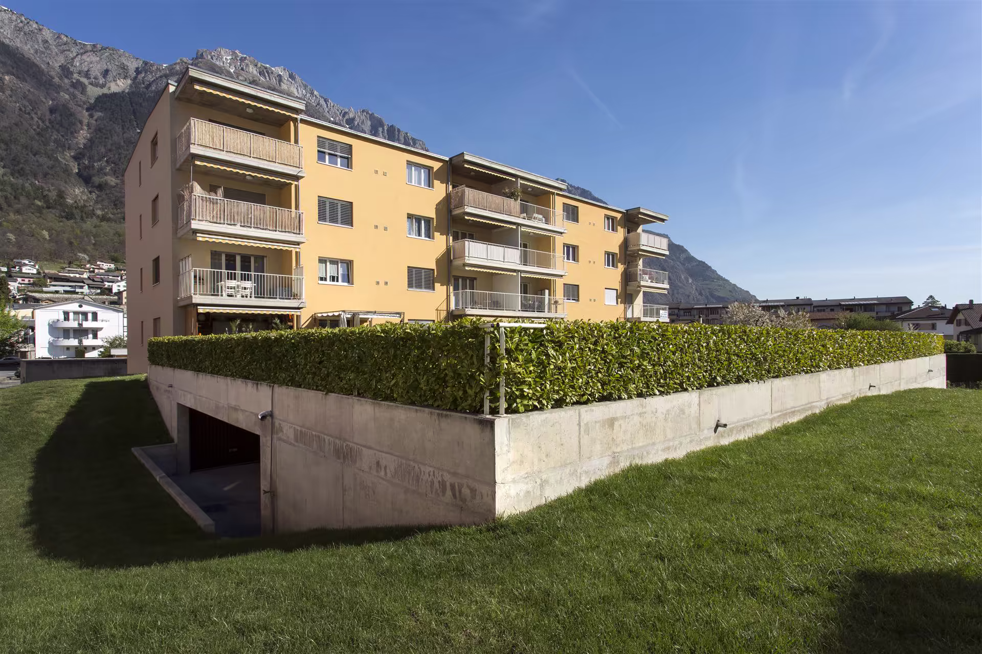 Immeuble résidentiel jaune avec balcons, haie verte et parking souterrain, situé au pied d’une montagne rocheuse sous un ciel bleu clair.
