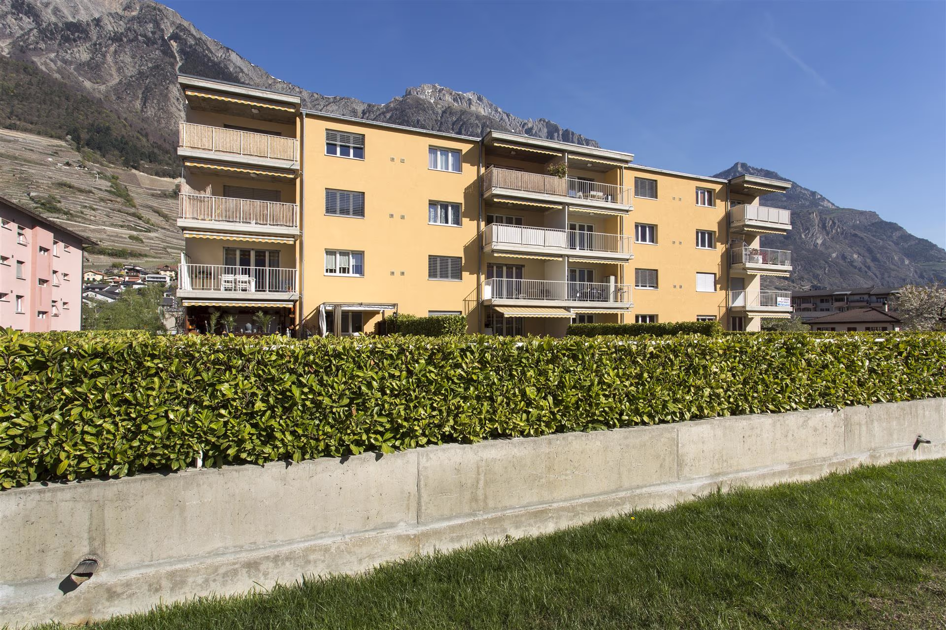 Immeuble jaune de quatre étages avec balcons, entouré de haies vertes, sur fond de montagnes rocheuses sous un ciel bleu clair.