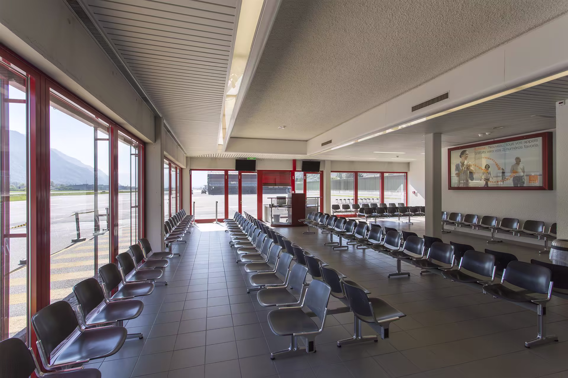 Salle d’attente d’aéroport vide avec rangées de sièges noirs, grandes baies vitrées rouges donnant sur la piste et panneaux publicitaires muraux.