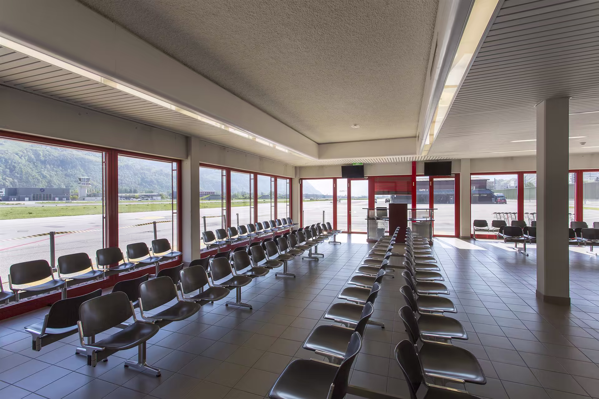 Salle d’attente d'aéroport vide avec rangées de sièges gris alignés, grandes fenêtres rouges donnant sur piste et hangars au loin.