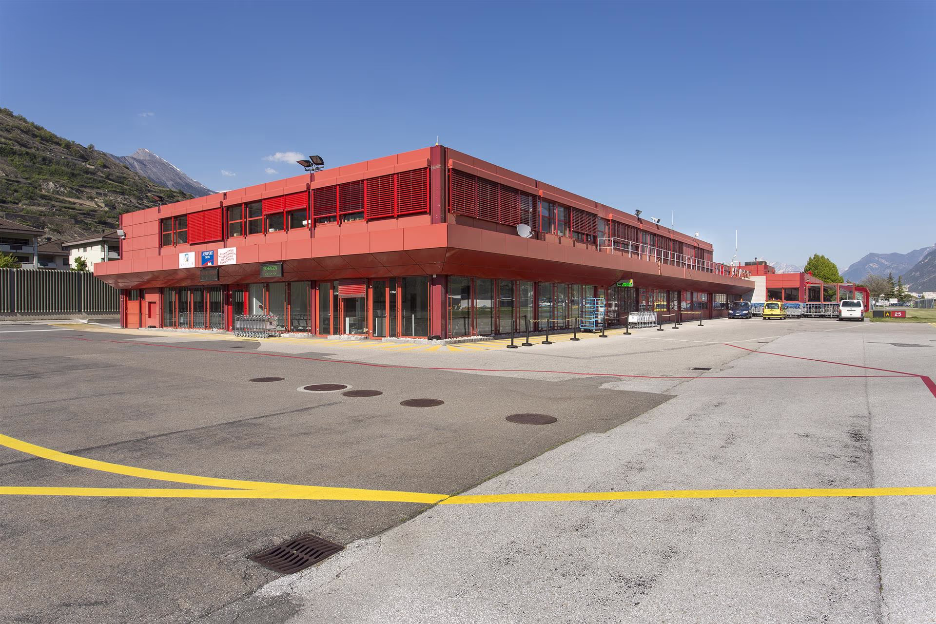 Bâtiment rouge moderne d'un terminal aéroportuaire avec parking, caddies et montagnes en arrière-plan sous un ciel clair.