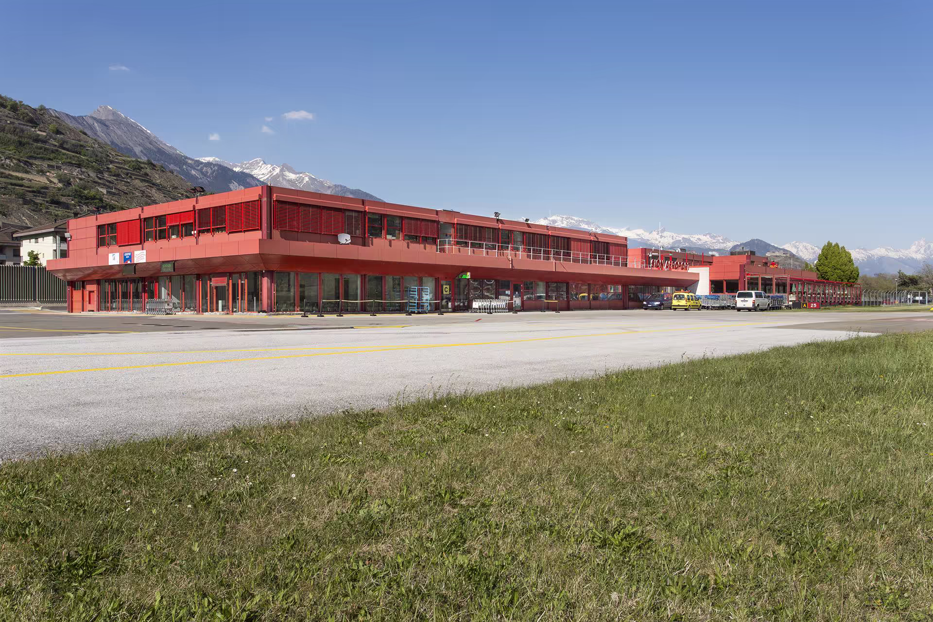 Terminal rouge d’un petit aéroport avec vue sur pistes, pelouse en premier plan et montagnes enneigées en arrière-plan sous ciel bleu.
