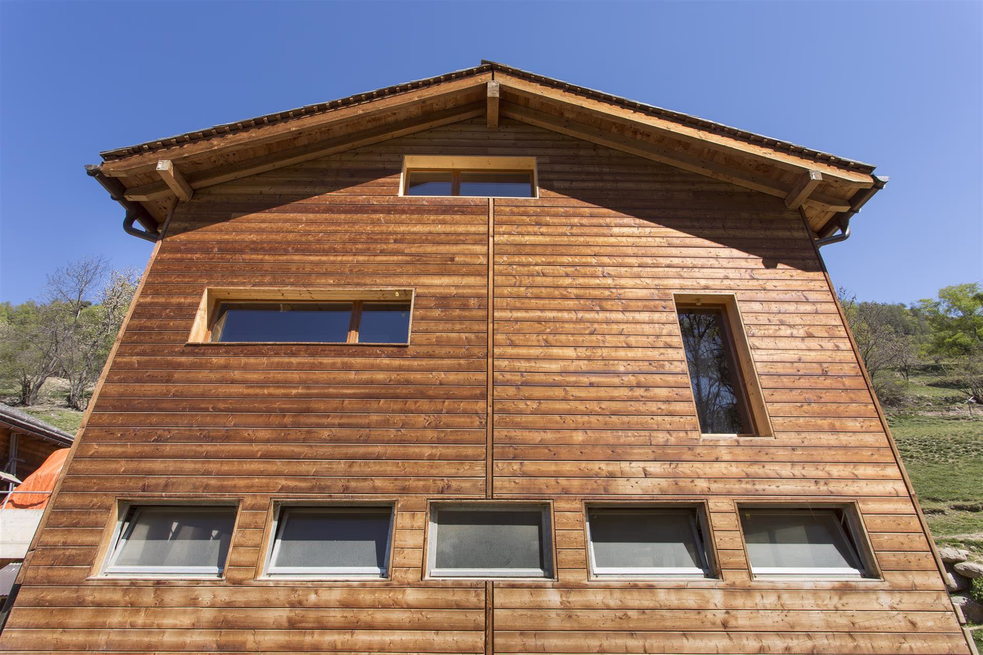 Façade en bois d’une maison avec sept fenêtres de différentes formes sous un toit à deux pans, ciel bleu et arbres au loin.