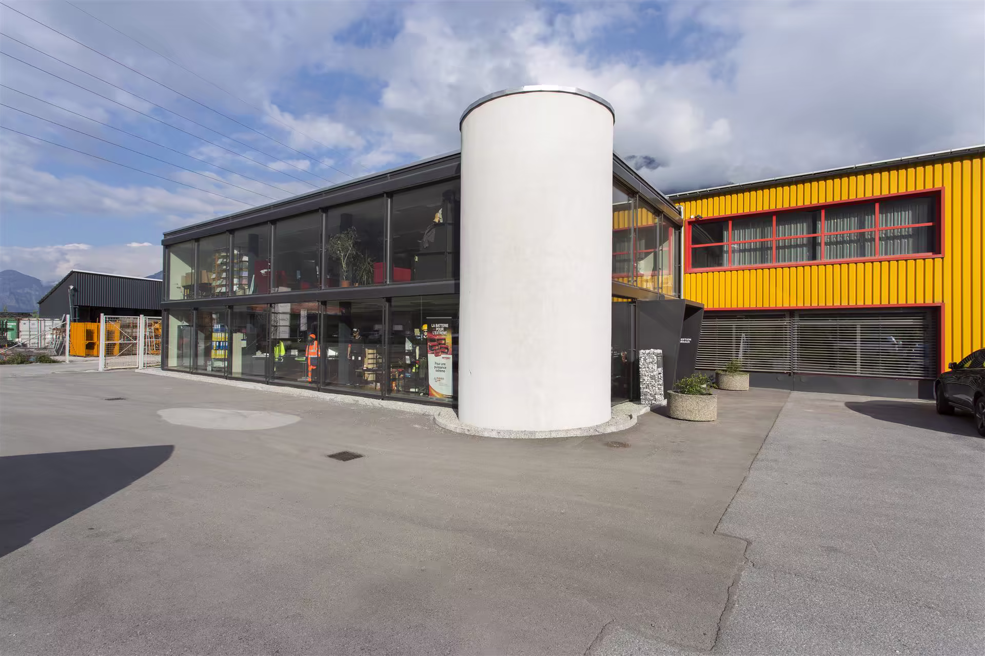Bâtiment industriel moderne avec murs vitrés, cylindre blanc central, façade jaune à fenêtres rouges, ciel nuageux et montagne au loin.