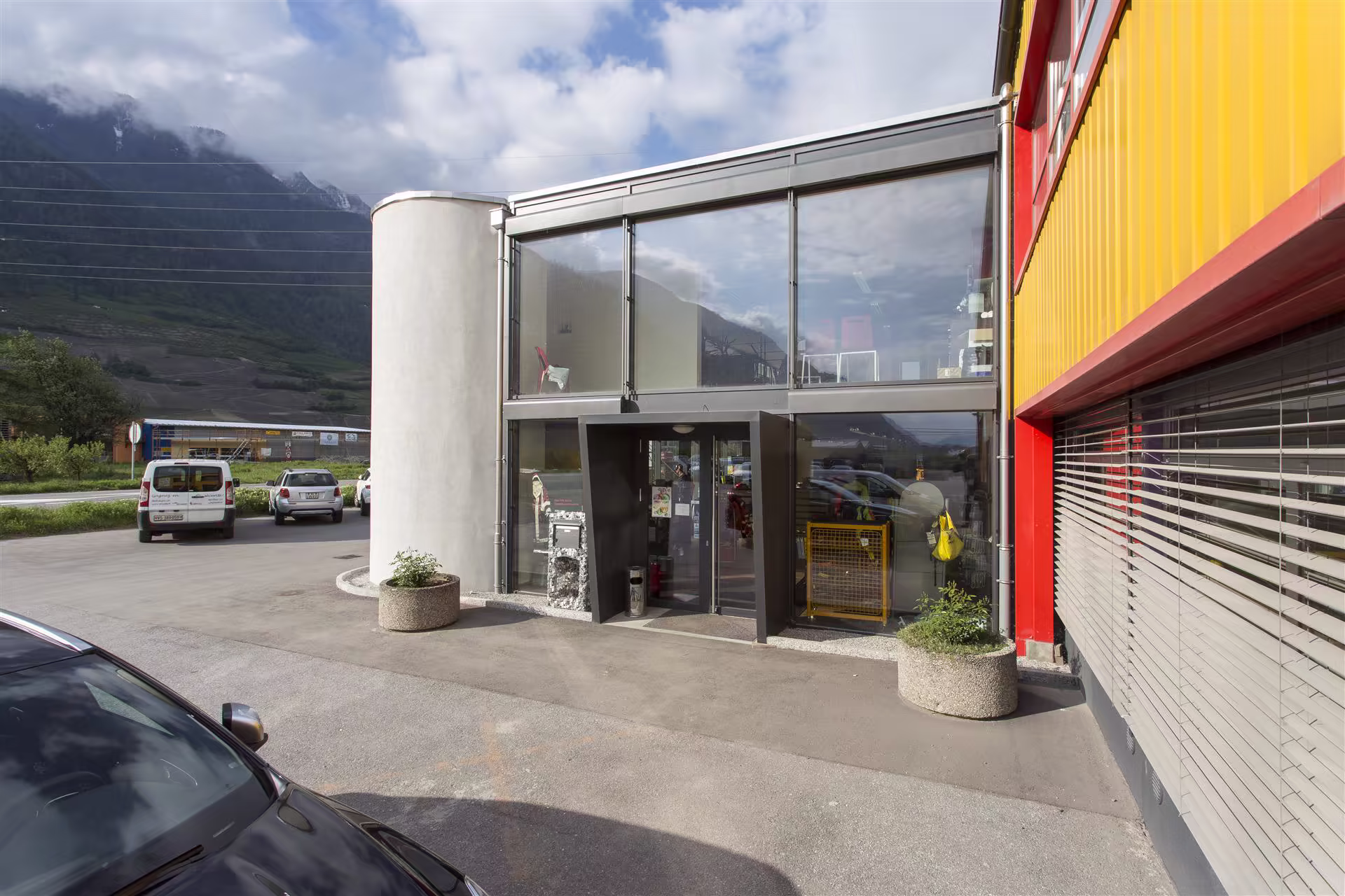 Façade d’un bâtiment industriel avec entrée vitrée, murs jaunes et rouges, parking et montagne en arrière-plan sous ciel nuageux.