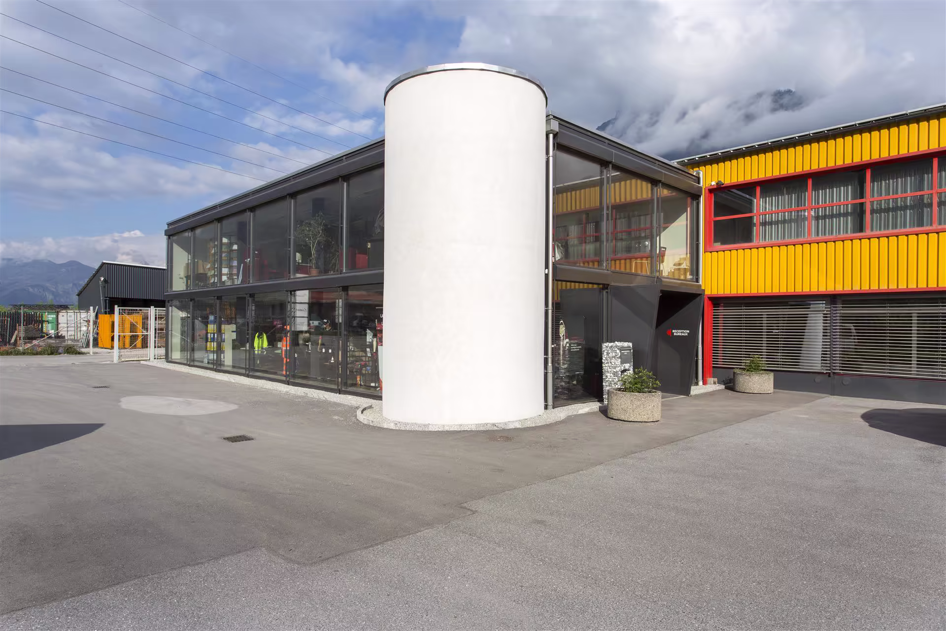 Bâtiment industriel moderne avec façade vitrée, cylindre blanc central, partie jaune à fenêtres rouges, deux grandes jardinières devant l'entrée.