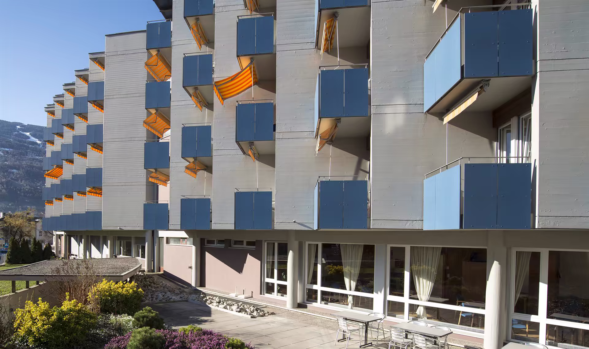 Façade d’un immeuble avec balcons bleus, stores orange et une terrasse au rez-de-chaussée, entourée de végétation et montagnes en arrière-plan.