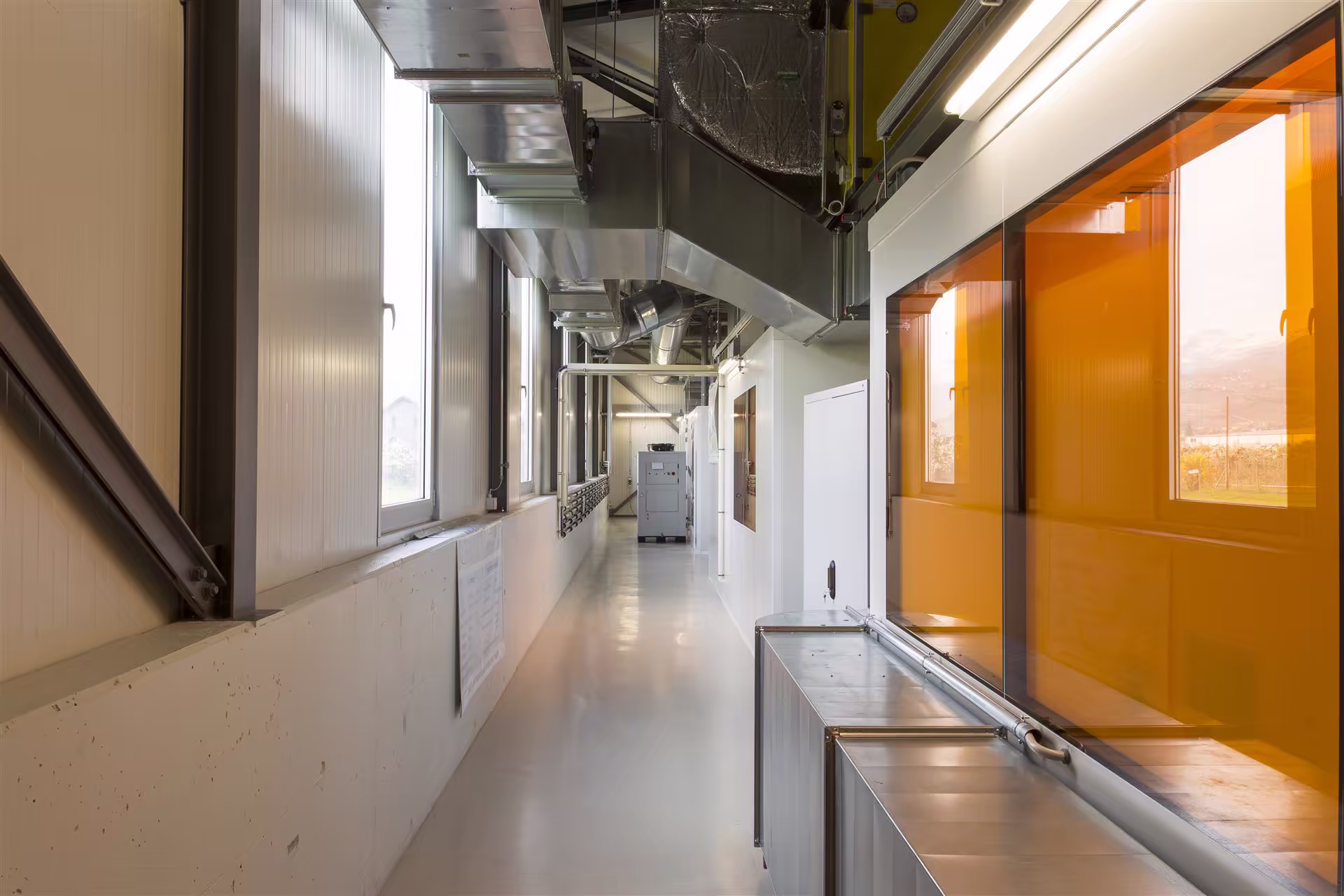 Couloir industriel avec grandes fenêtres, conduits métalliques au plafond, mur avec panneaux blancs et vitres teintées orange sur la droite.
