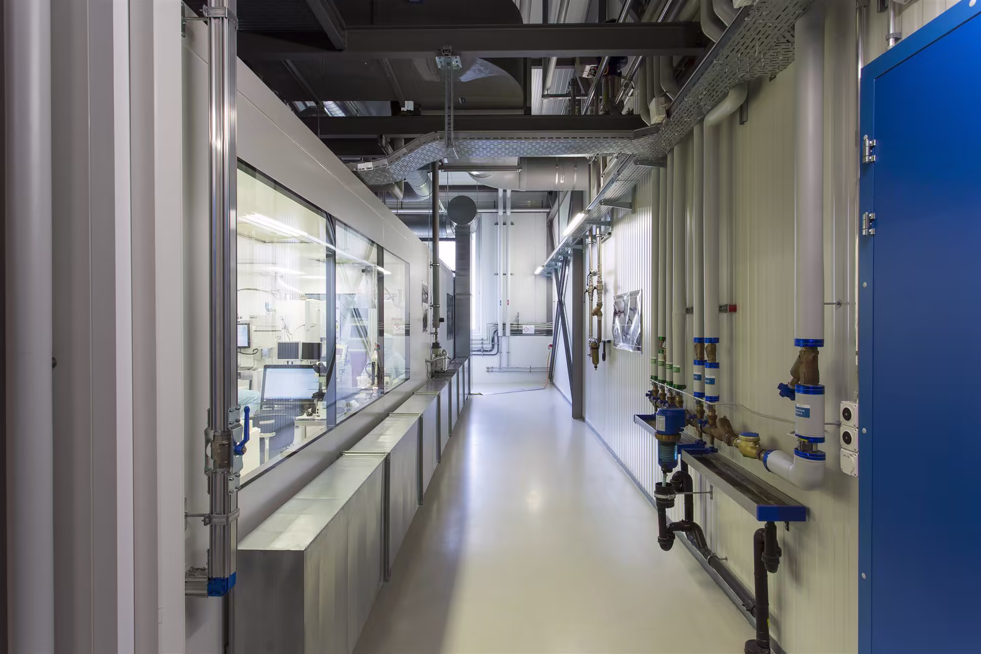 Couloir industriel propre avec tuyauterie bleue et blanche sur le mur droit, baie vitrée à gauche donnant sur une salle de contrôle avec ordinateurs.