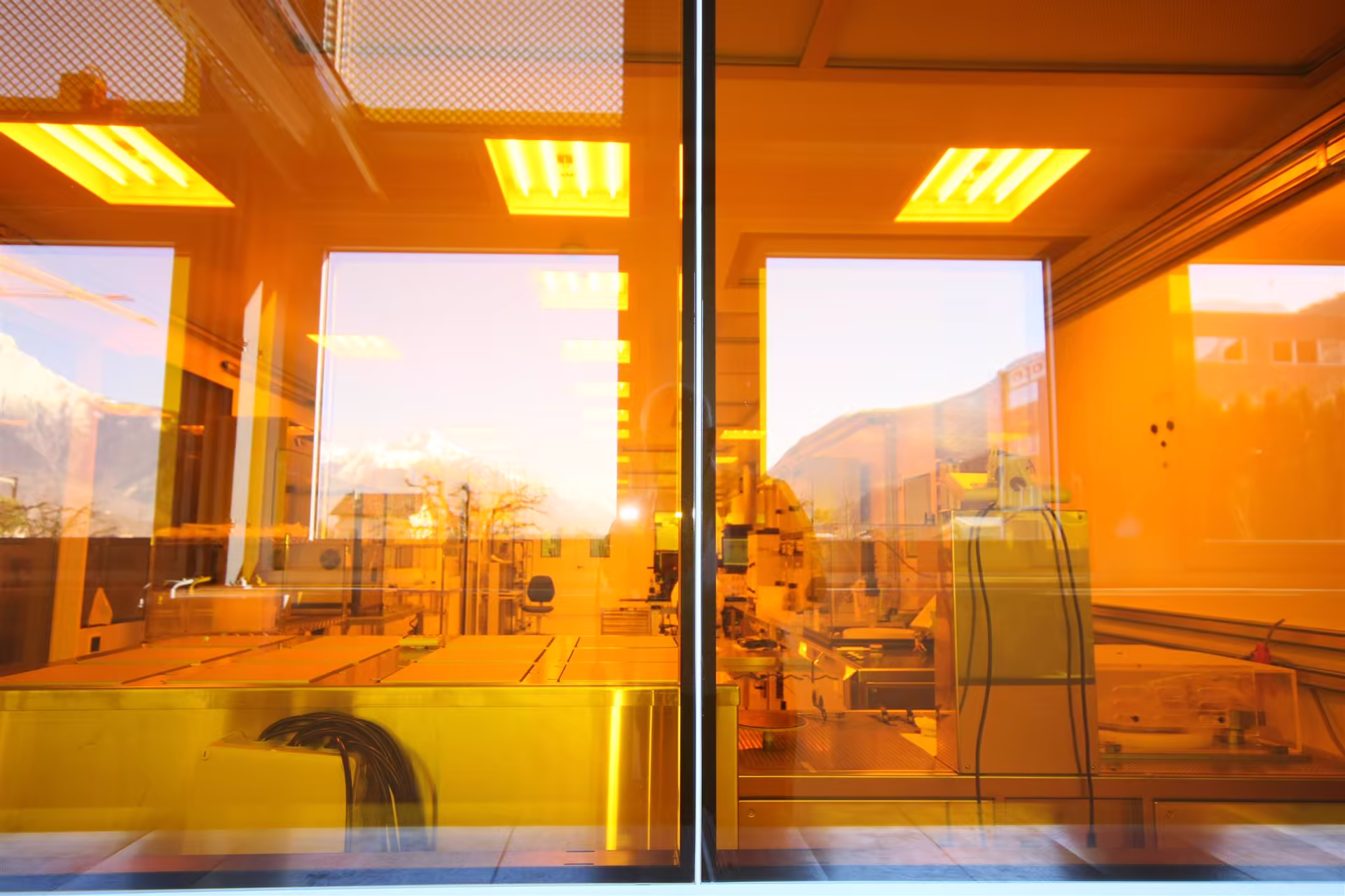 Intérieur d'un laboratoire ou salle blanche visible à travers une vitre teintée orange, avec équipement technique et reflets de montagnes à l'extérieur.