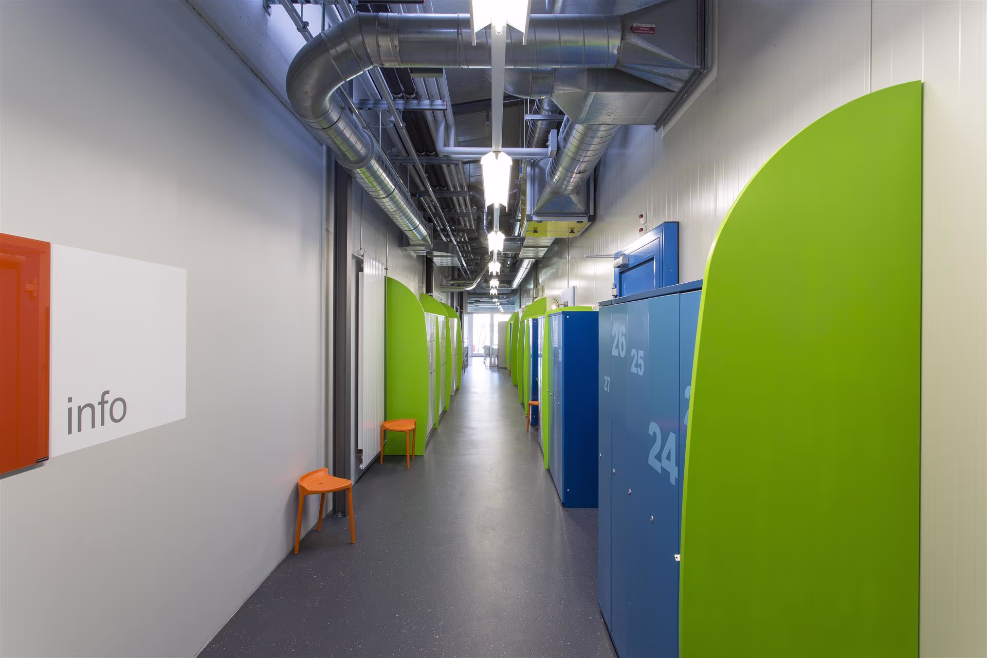 Couloir industriel éclairé avec casiers bleus numérotés, séparateurs verts, tabourets orange et conduits d’aération métalliques apparents au plafond.