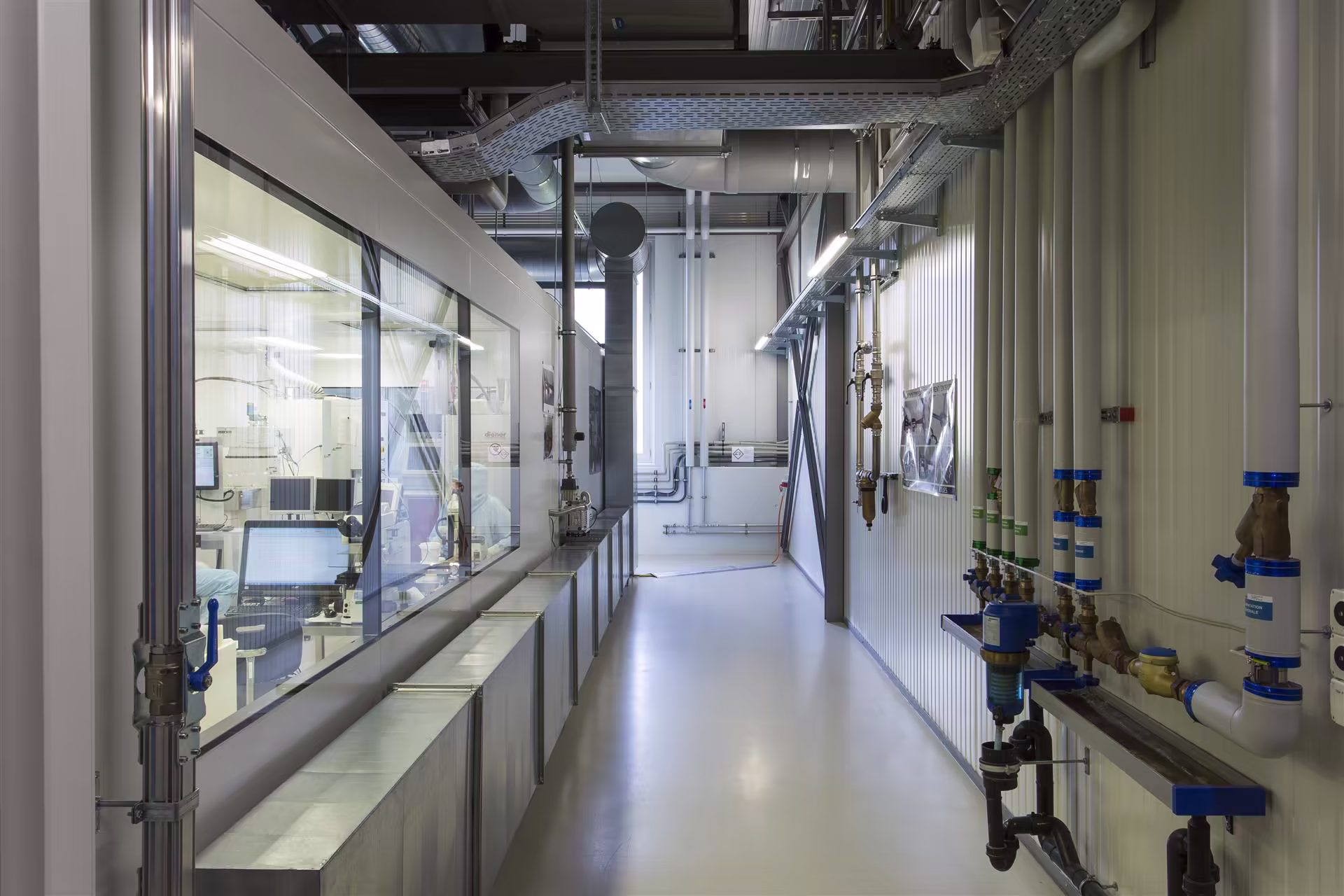 Couloir industriel propre avec gaines d’aération et plomberie sur les murs, fenêtres donnant sur une salle de laboratoire avec équipements et personnel en tenue blanche.