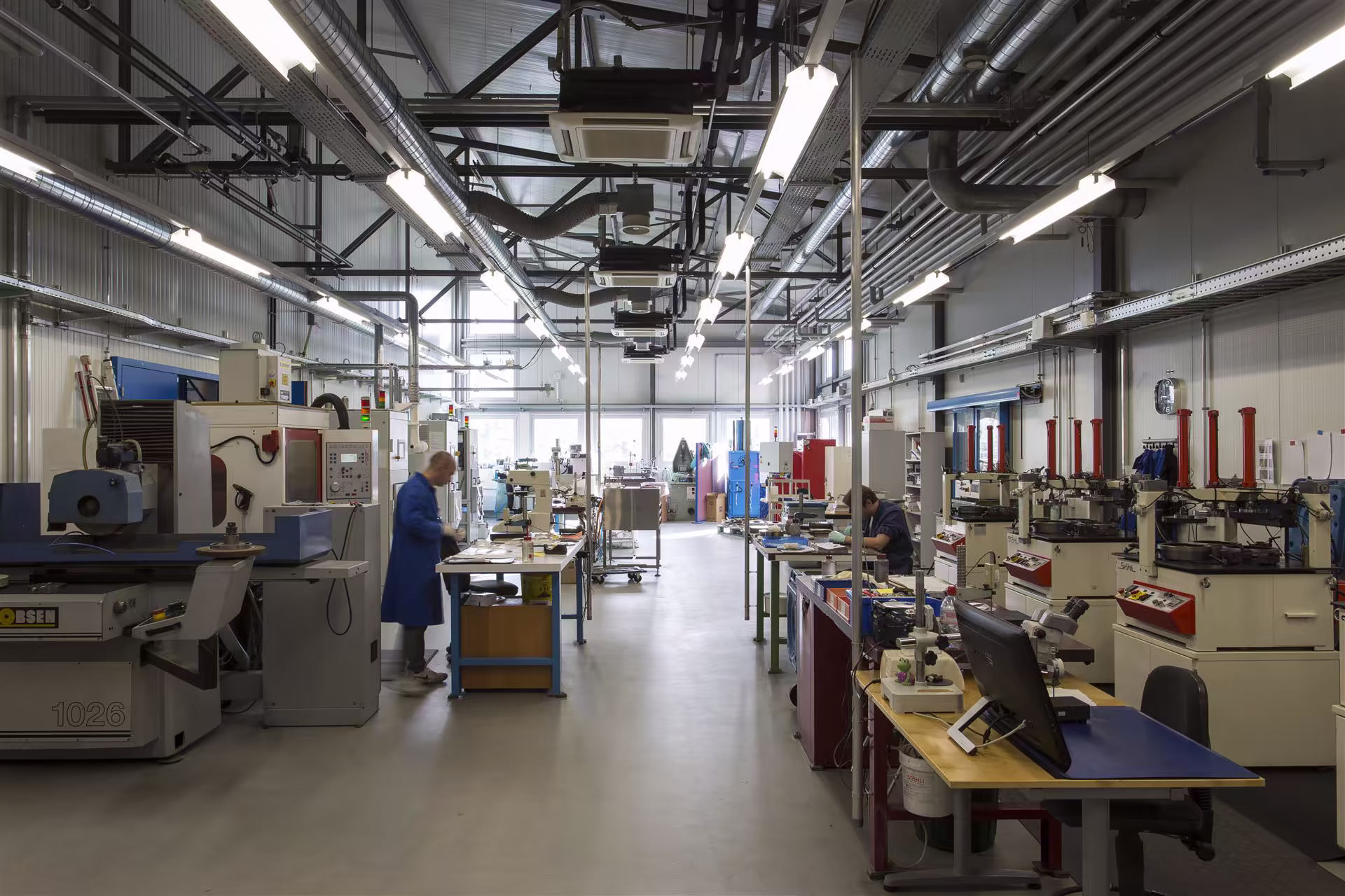 Atelier industriel lumineux avec machines-outils, deux ouvriers en train de travailler à leurs postes, nombreux équipements et tables de travail.