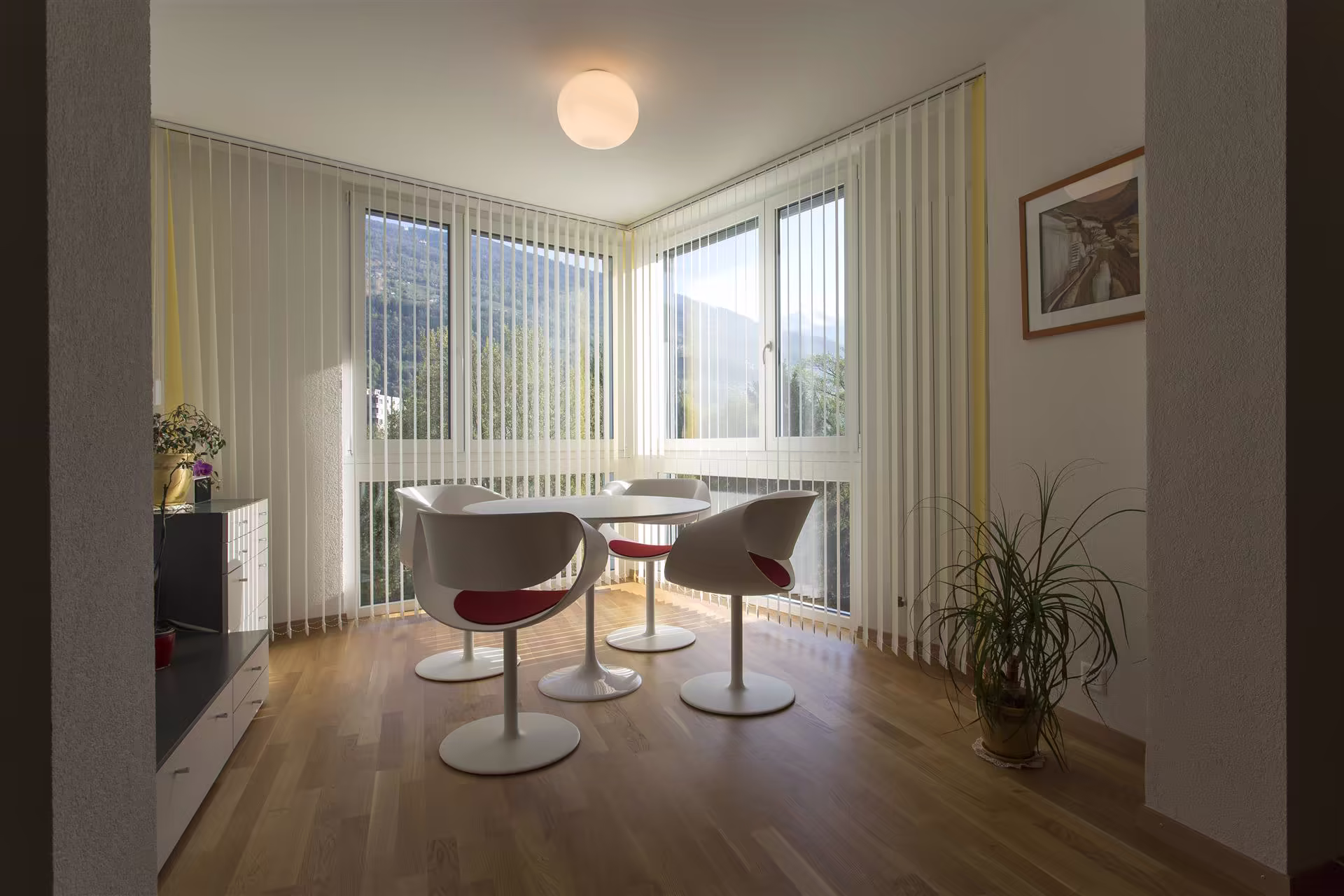 Coin repas lumineux avec table ronde blanche, quatre chaises blanches à coussins rouges, fenêtres à stores verticaux et vue sur montagnes verdoyantes.