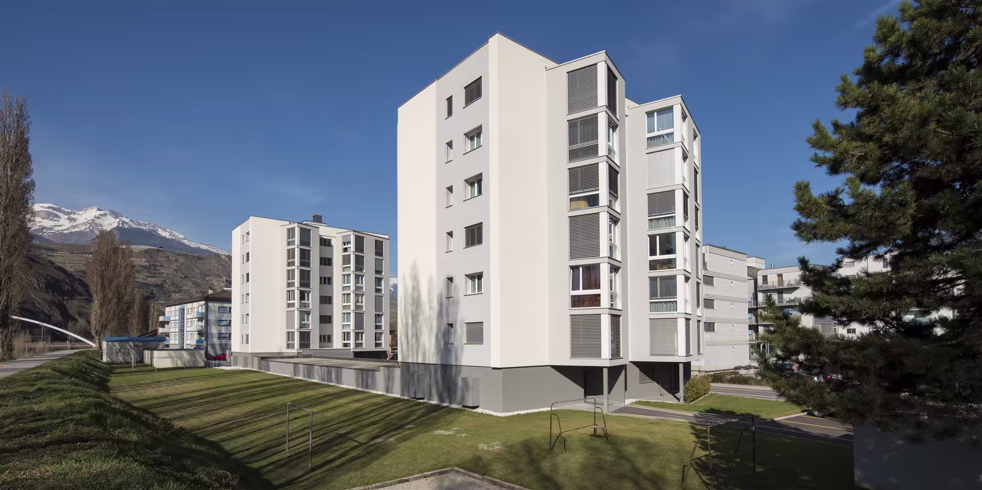 Deux immeubles blancs modernes de plusieurs étages entourés d’un espace vert avec montagnes enneigées en arrière-plan sous ciel bleu.