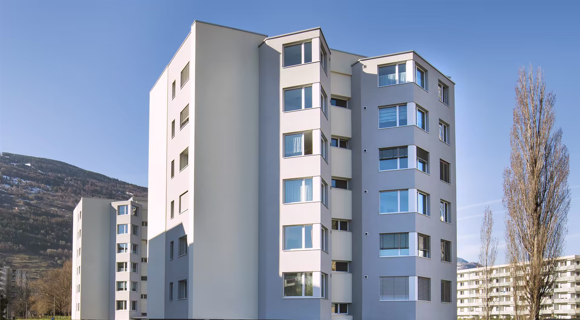 Immeubles résidentiels modernes et blancs sous un ciel bleu, avec des arbres en arrière-plan et une montagne partiellement enneigée.