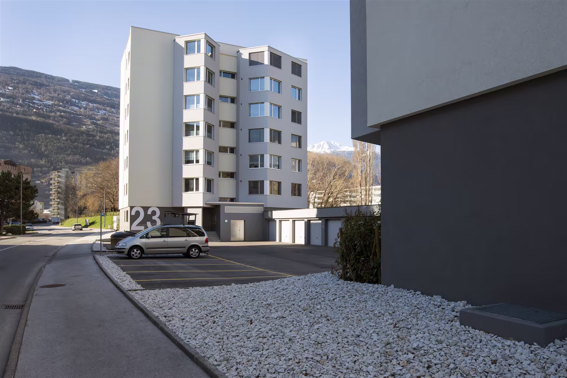 Bâtiment résidentiel gris clair avec le numéro 23, un parking avec une voiture grise, des garages blancs, montagnes enneigées en arrière-plan.