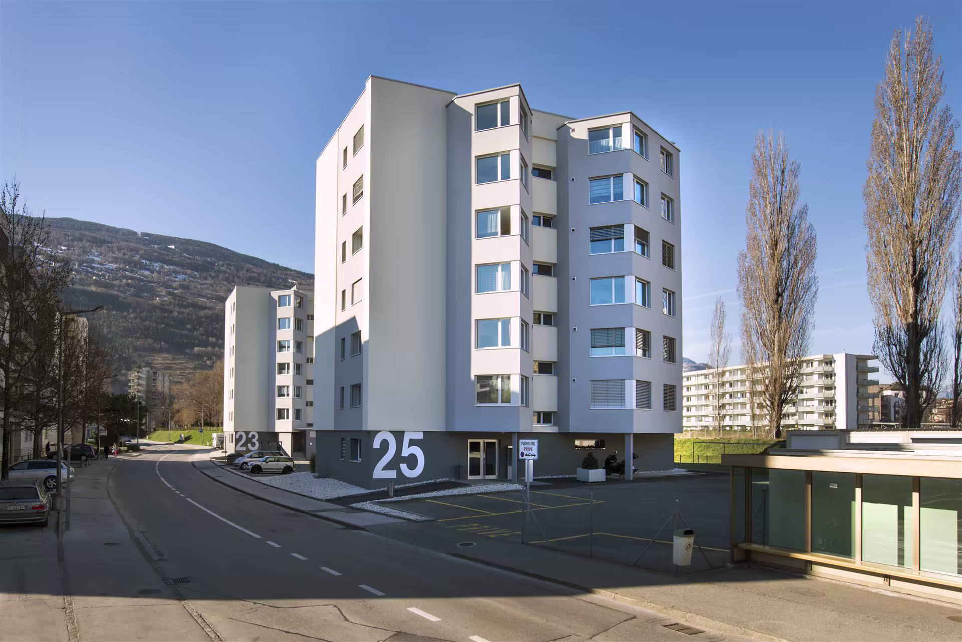 Immeubles résidentiels modernes numérotés 23 et 25, route courbe, voitures stationnées, arbres dépouillés, montagne en arrière-plan sous ciel bleu clair.