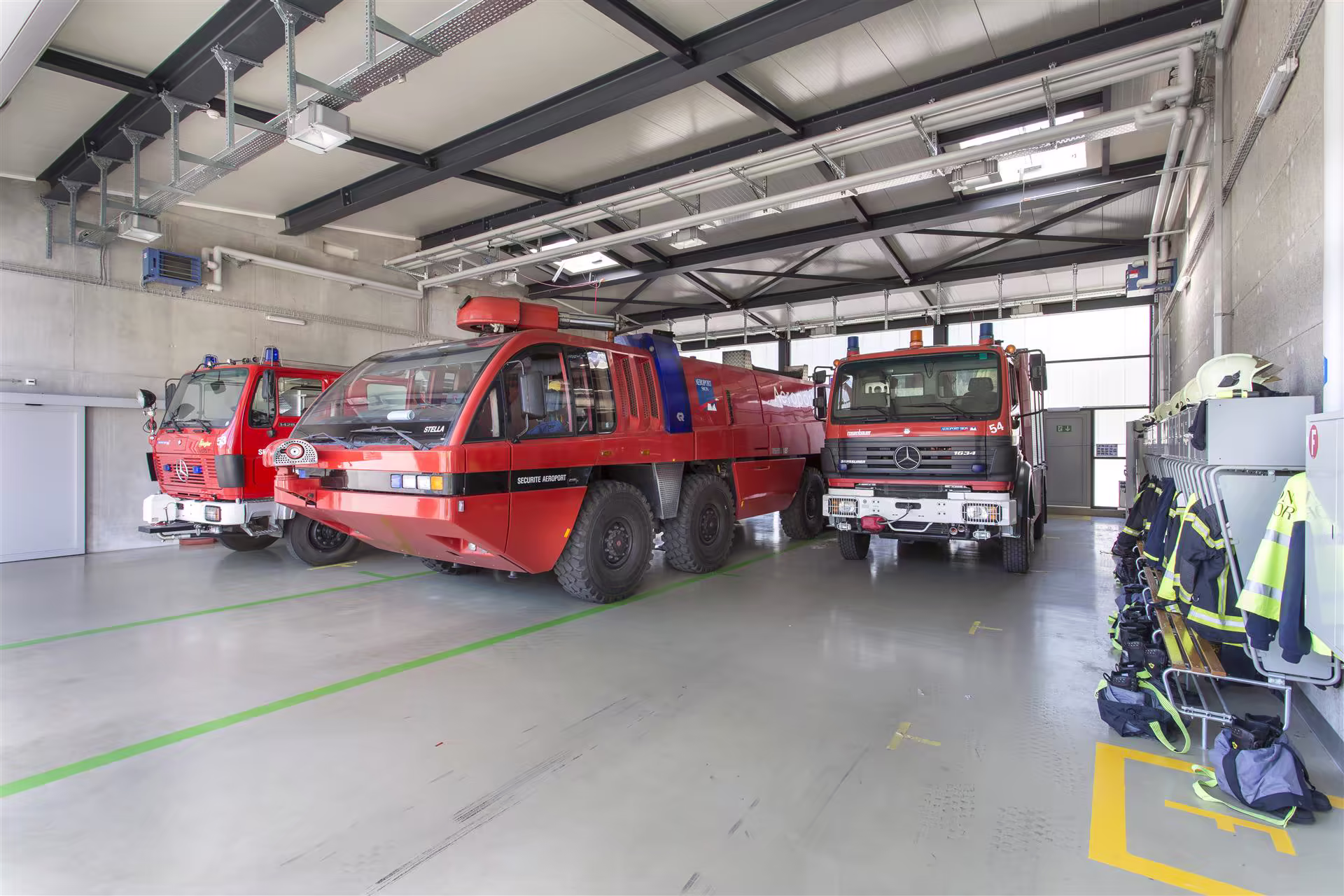 Trois camions de pompiers rouges stationnés dans un garage, avec des tenues et casques suspendus sur des crochets à droite.