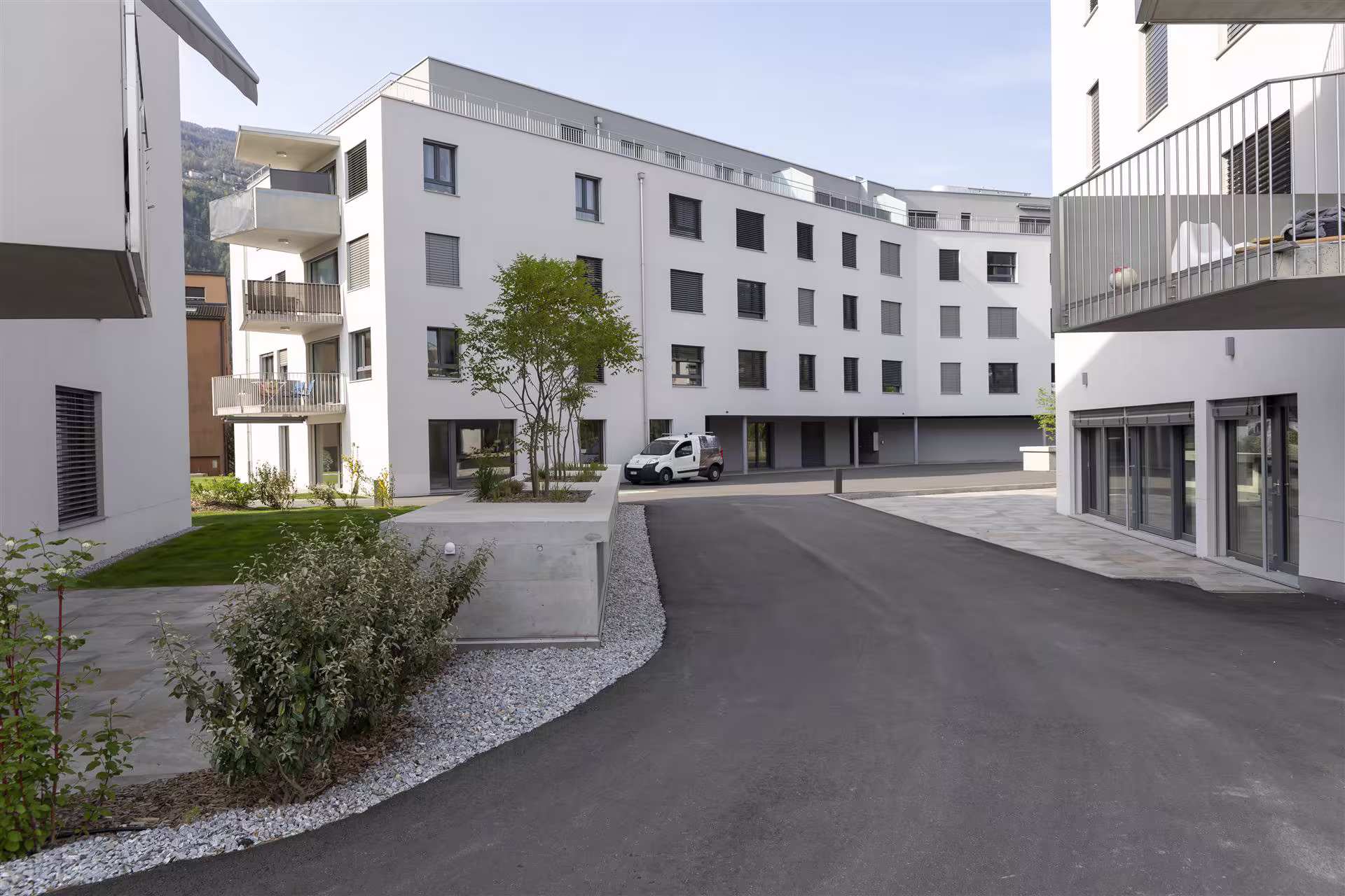 Cour intérieure d’un complexe résidentiel moderne avec plusieurs bâtiments blancs, balcons, espaces verts, et une voiture blanche stationnée.