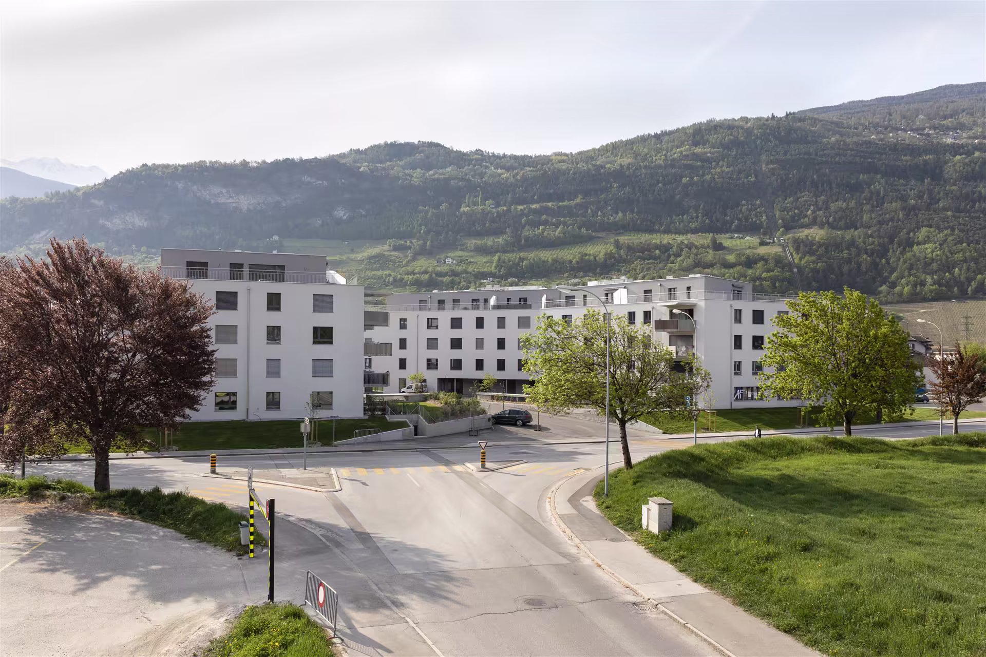 Quartier résidentiel avec immeubles blancs modernes, arbres verts et rouges, routes vides, pelouses et montagnes boisées en arrière-plan sous ciel clair.
